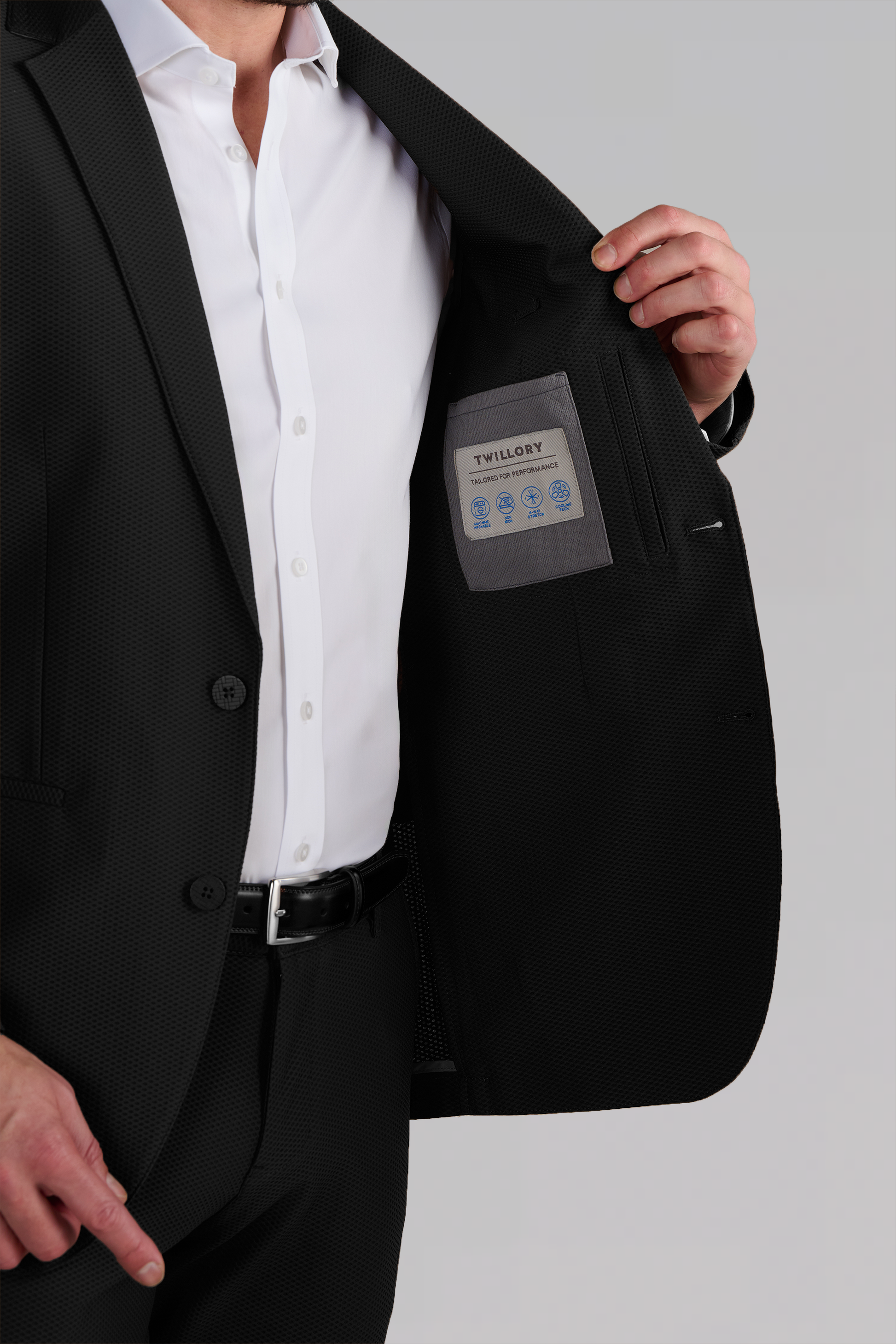 A man in a black Twillory AIR Blazer holds open his jacket to reveal the inner pocket with care symbols and a label. He wears a white shirt, his head isn't visible, and the background is plain light gray.