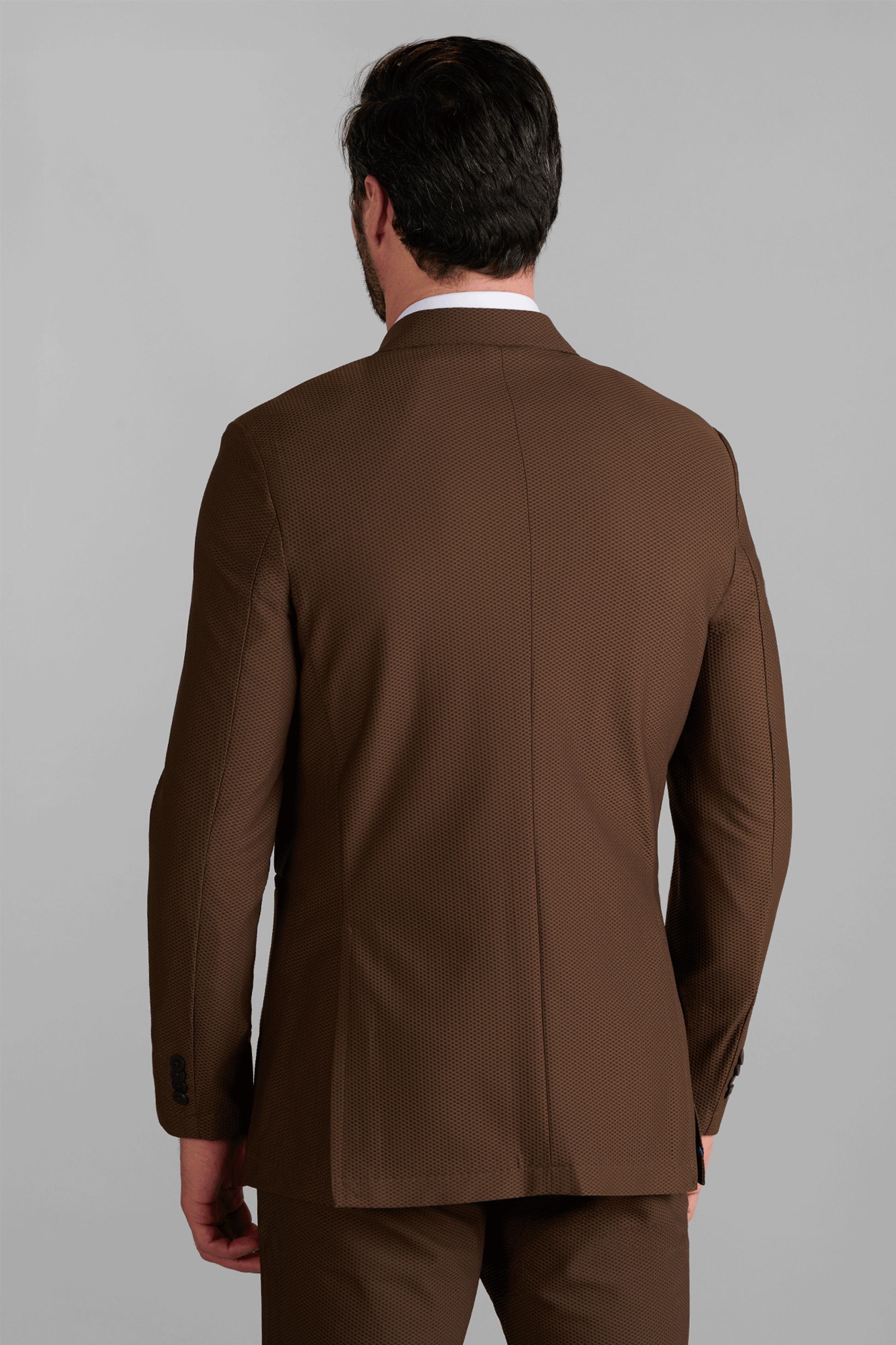 A man with short dark hair is seen from behind, wearing the Twillory AIR Blazer // Brown and matching trousers. He stands against a plain gray background; the blazer features subtle texture and visible seams.