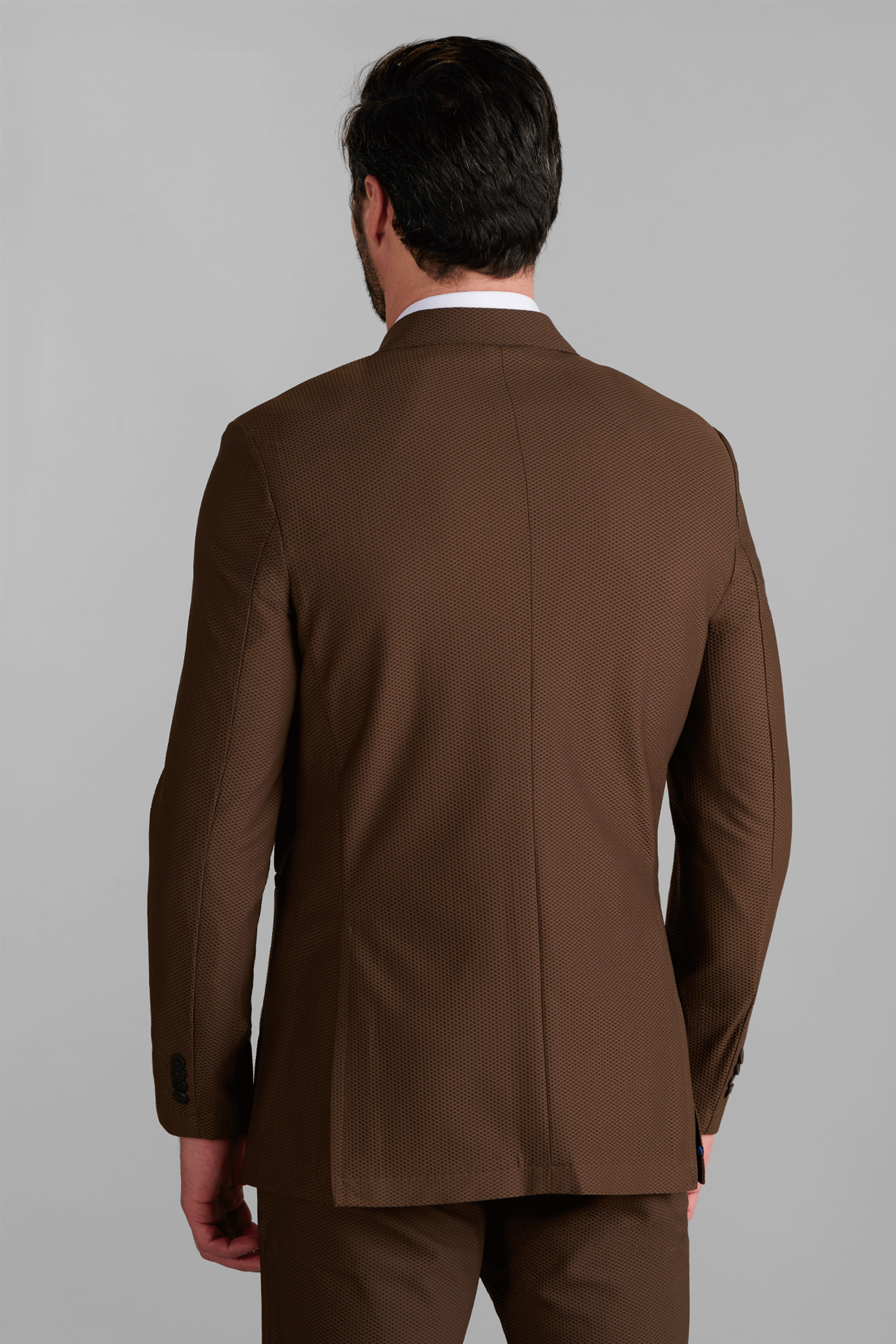 A man with short dark hair is seen from behind, wearing the Twillory AIR Blazer // Brown and matching trousers. He stands against a plain gray background; the blazer features subtle texture and visible seams.