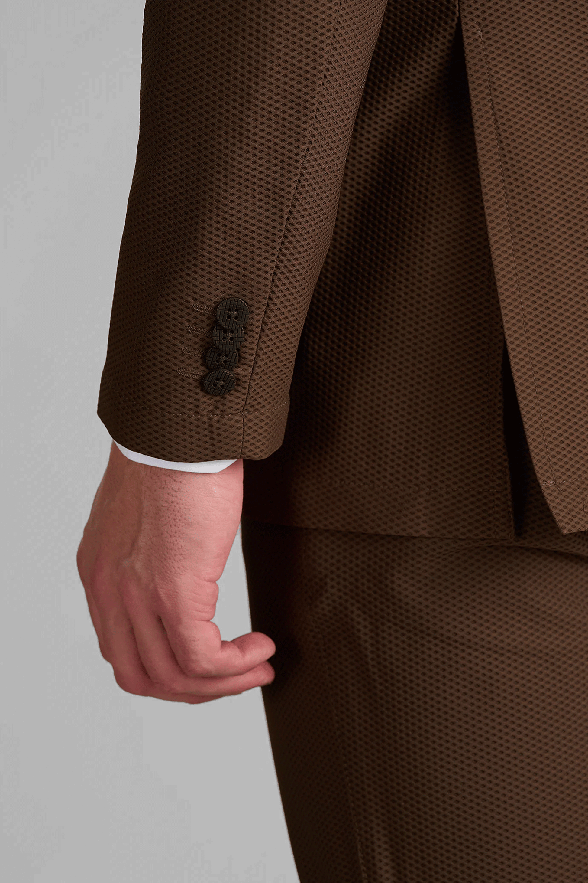 A close-up of a person’s left arm and hip, wearing the Twillory AIR Blazer // Brown. This machine-washable blazer features breathable performance fabric with black sleeve buttons, and a white shirt cuff is visible at the wrist.
