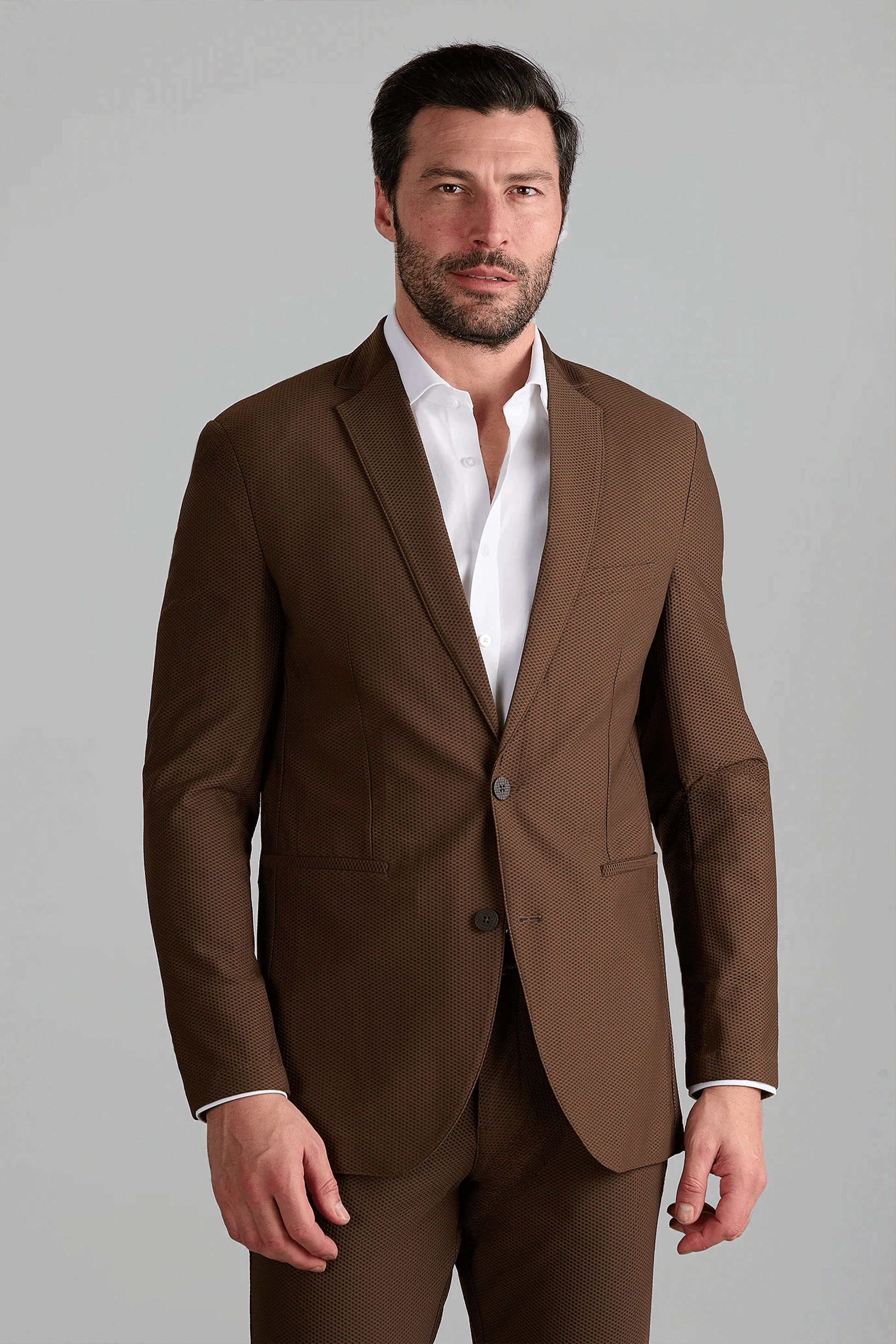 A man with short dark hair and a trimmed beard stands against a gray background, wearing the Twillory AIR Blazer in brown with matching trousers over a white dress shirt. His expression is neutral, left hand by his side.