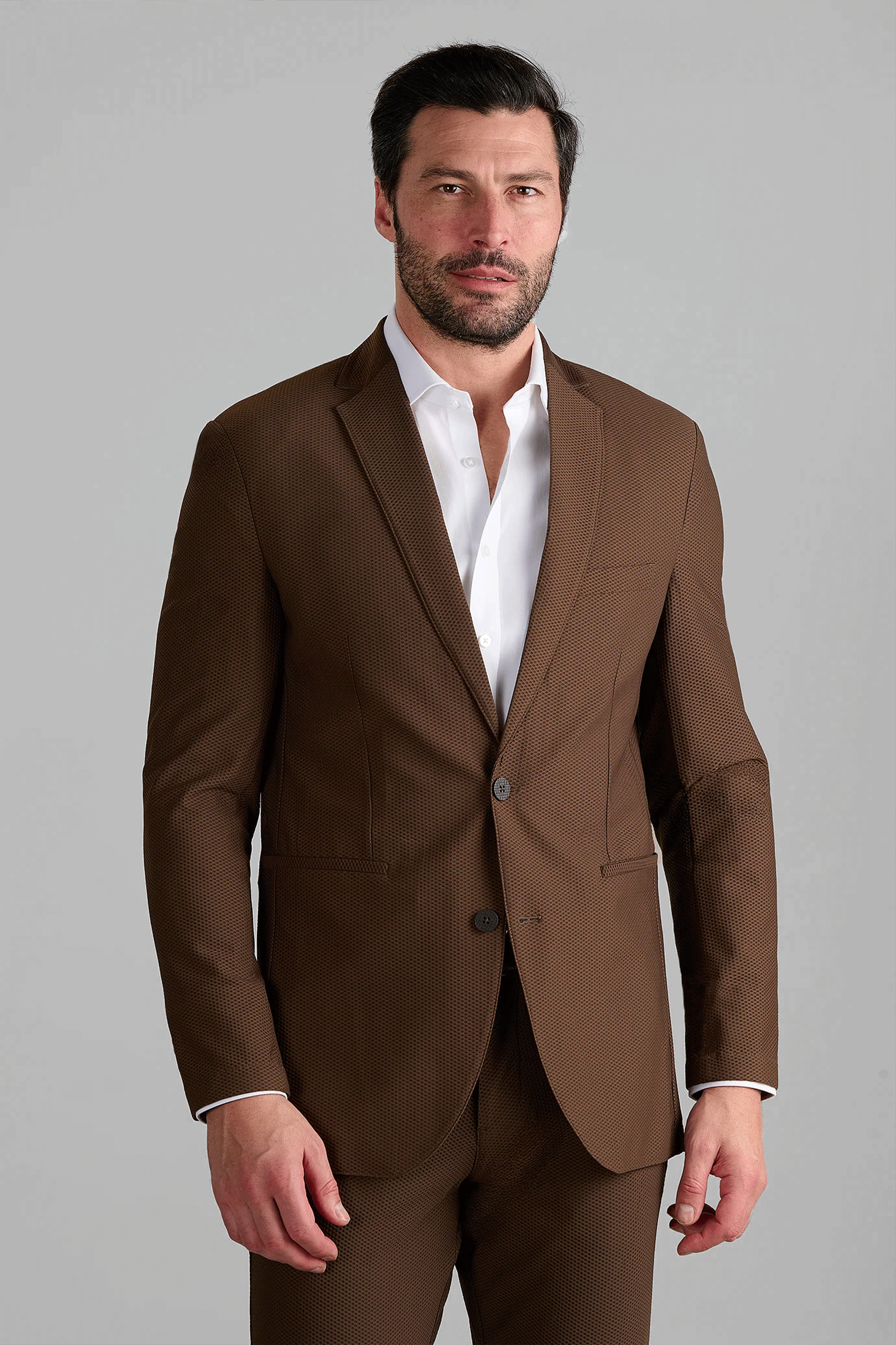 A man with short dark hair and a trimmed beard stands against a gray background, wearing the Twillory AIR Blazer in brown with matching trousers over a white dress shirt. His expression is neutral, left hand by his side.