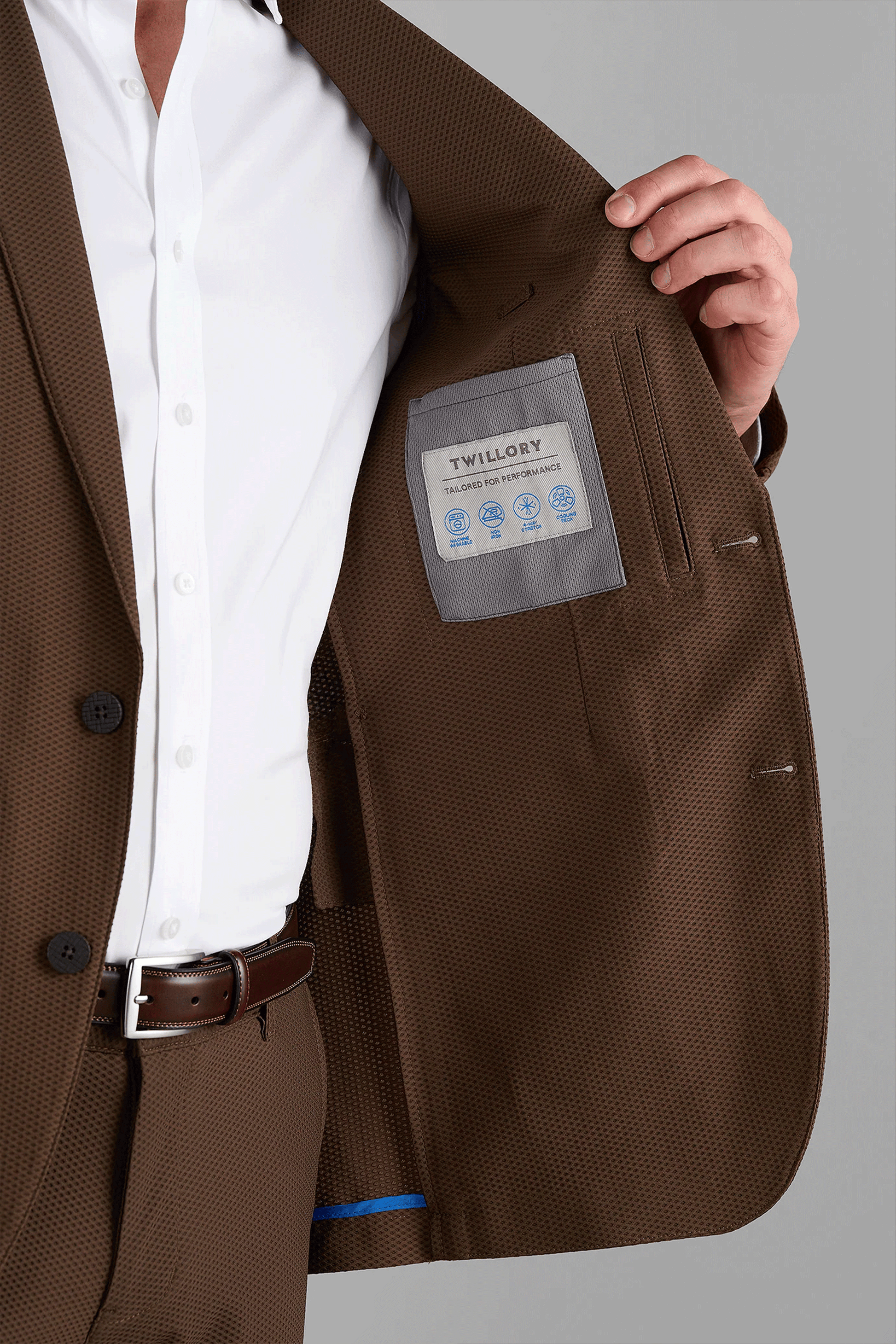 A person in a brown AIR Blazer by Twillory opens the jacket to reveal the label with care icons. The focus is on the machine washable suit’s lining and label; their head is out of frame.