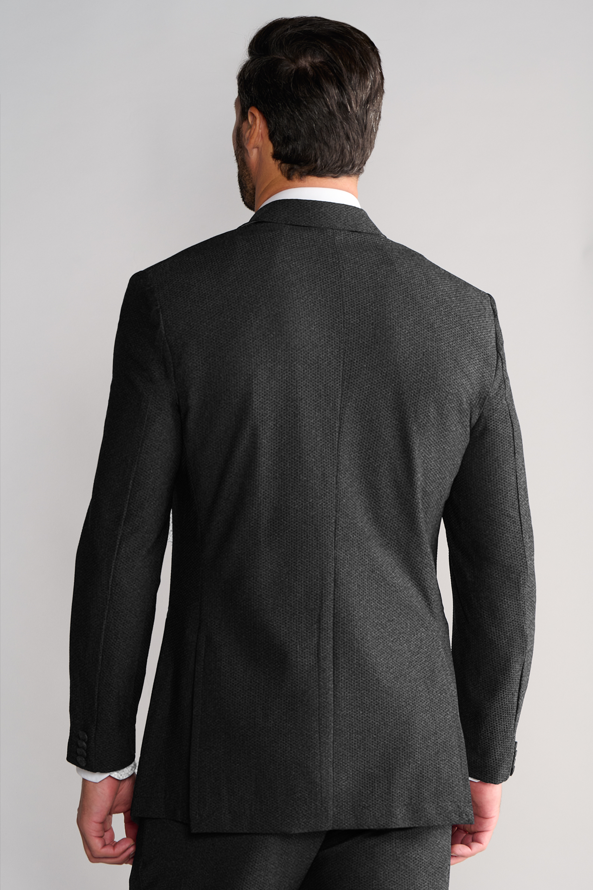 A man with short dark hair is seen from behind wearing Twillory’s AIR Blazer // Heather Grey, made with 360° airflow fabric. He stands naturally against a plain light gray background, a white shirt collar visible above the blazer.