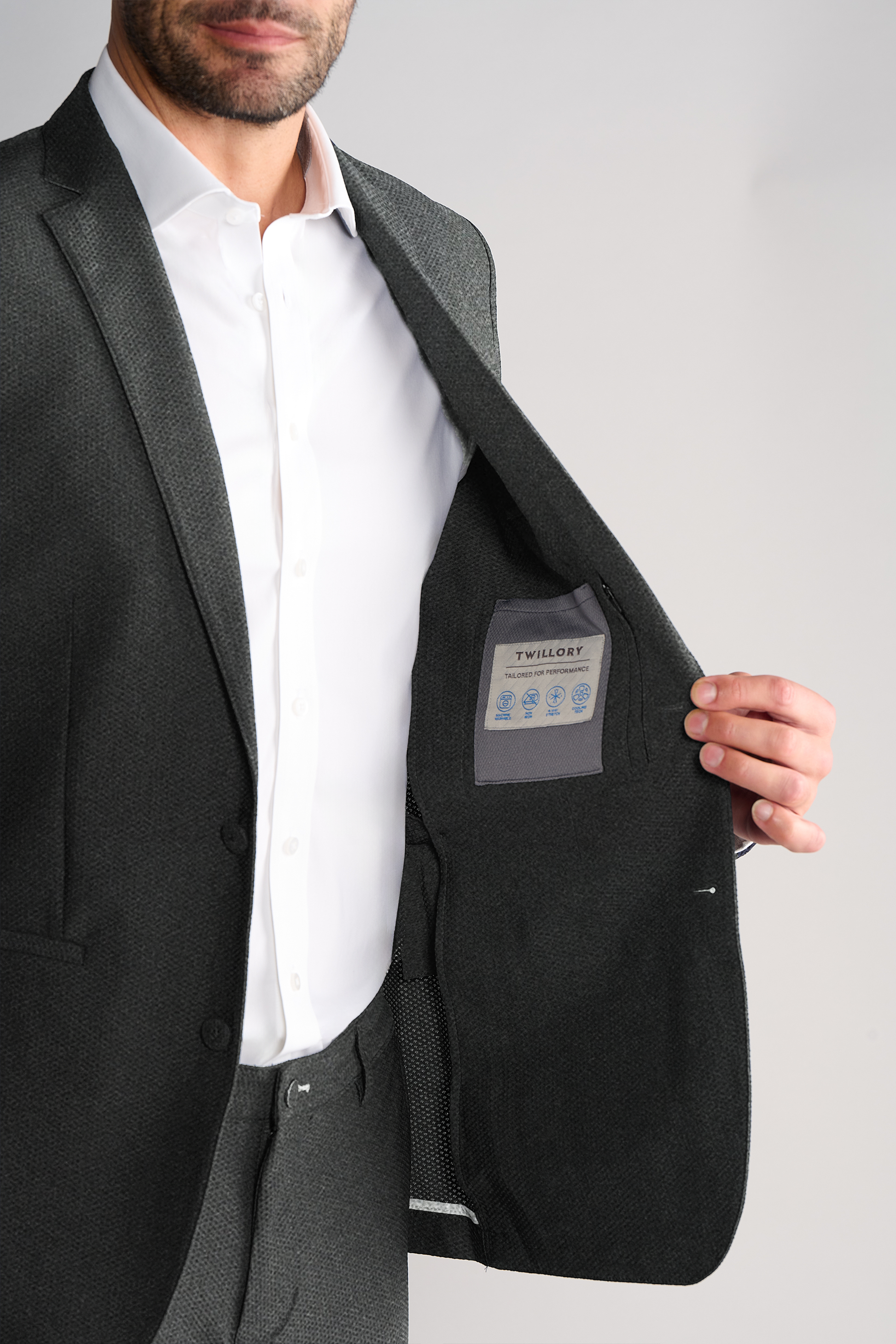 A man in a heather grey AIR Blazer by Twillory and a white dress shirt opens his machine-washable jacket to reveal the inside label with care symbols and Twillory branding, standing against a light gray background.