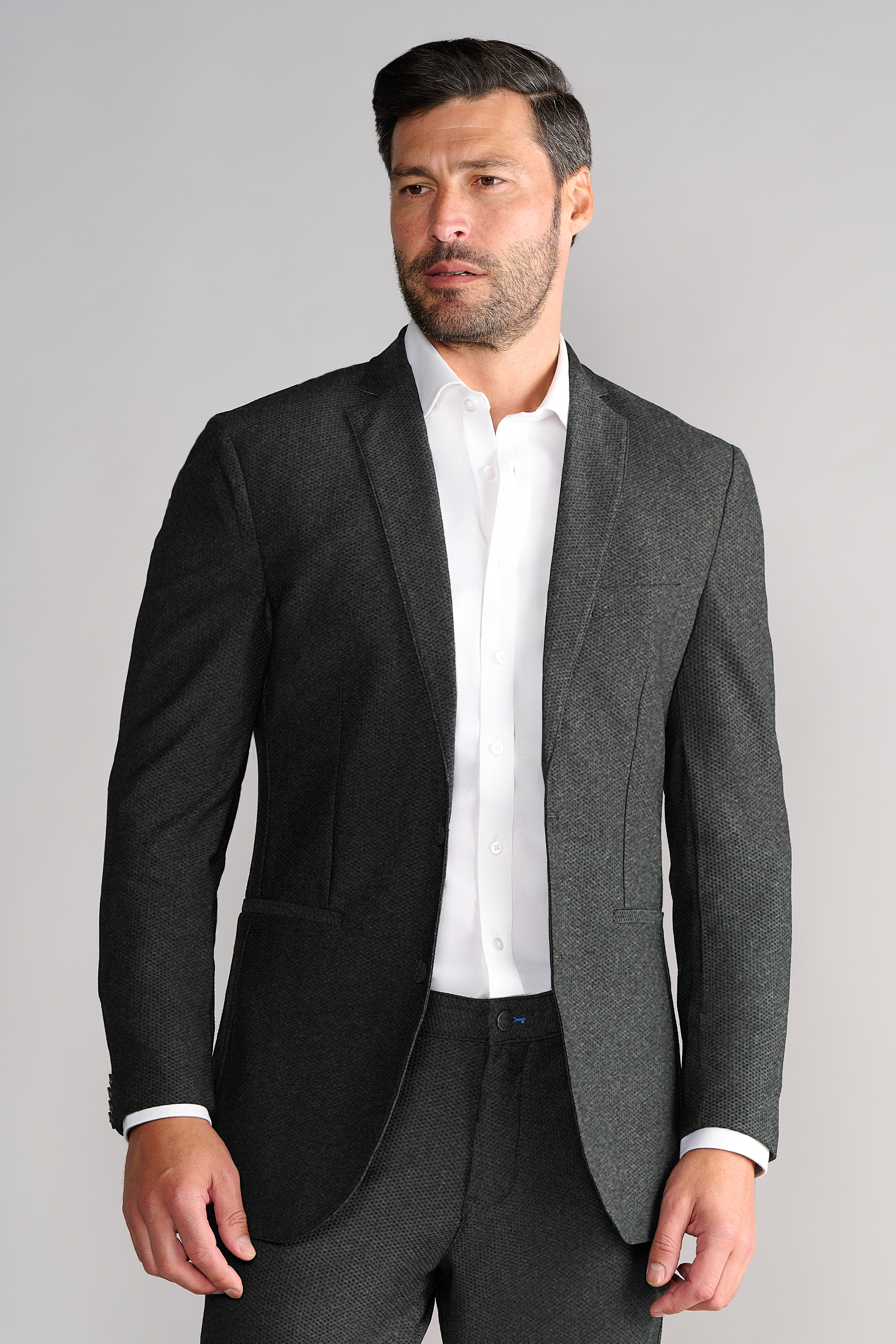 A man with short dark hair and a trimmed beard wears the Twillory AIR Blazer in Heather Grey over a white dress shirt, paired with matching pants. He faces slightly left against a plain light grey background, looking off-camera.