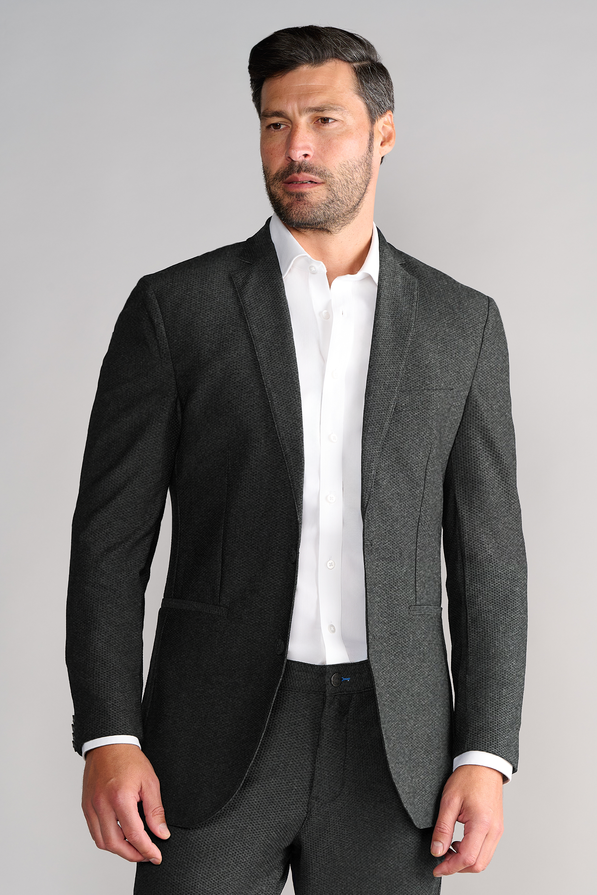 A man with short dark hair and a trimmed beard wears the Twillory AIR Blazer in Heather Grey over a white dress shirt, paired with matching pants. He faces slightly left against a plain light grey background, looking off-camera.