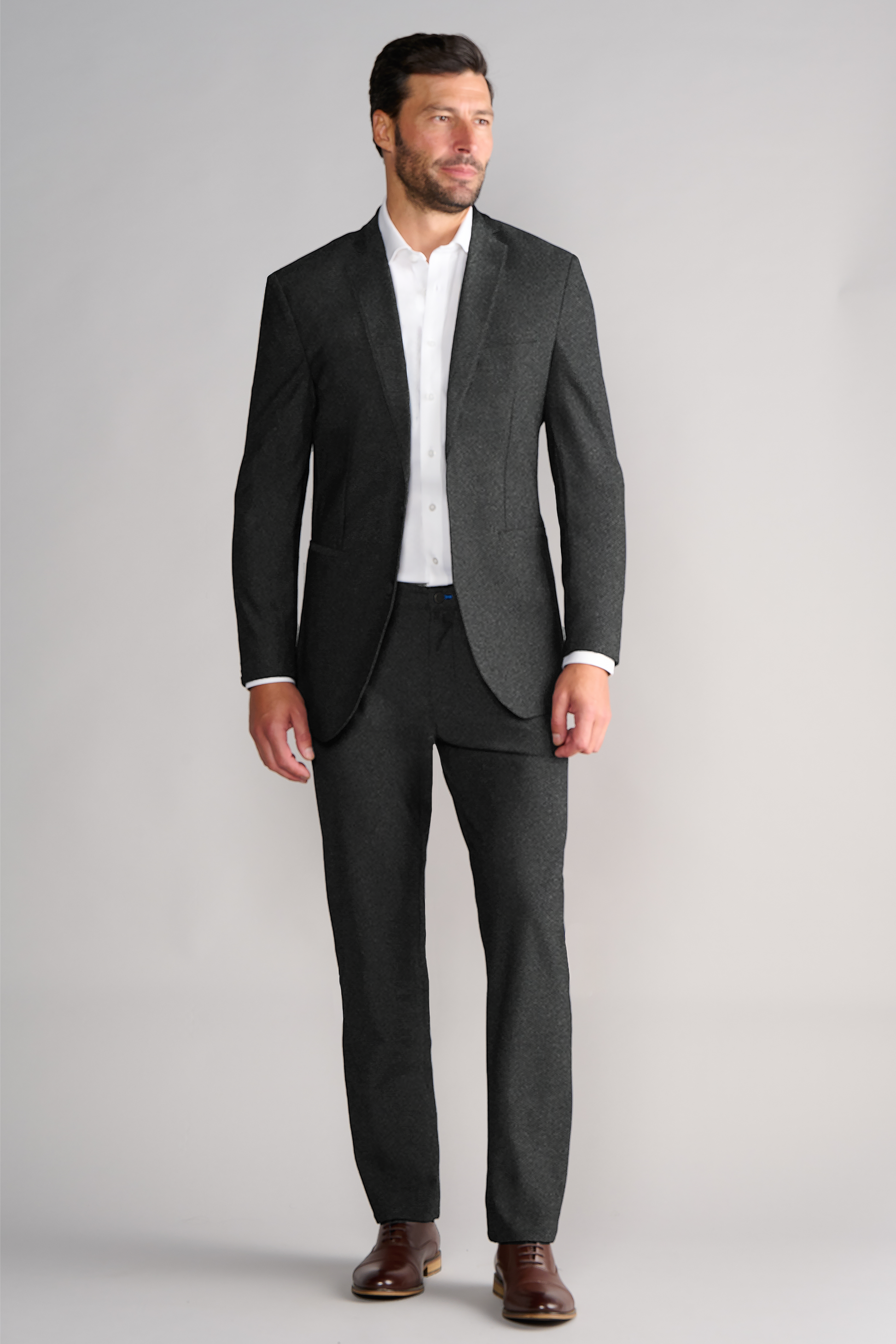 A man with short dark hair and a trimmed beard stands confidently against a light gray background, wearing Twillory’s AIR Blazer // Heather Grey over a white dress shirt and brown leather shoes.