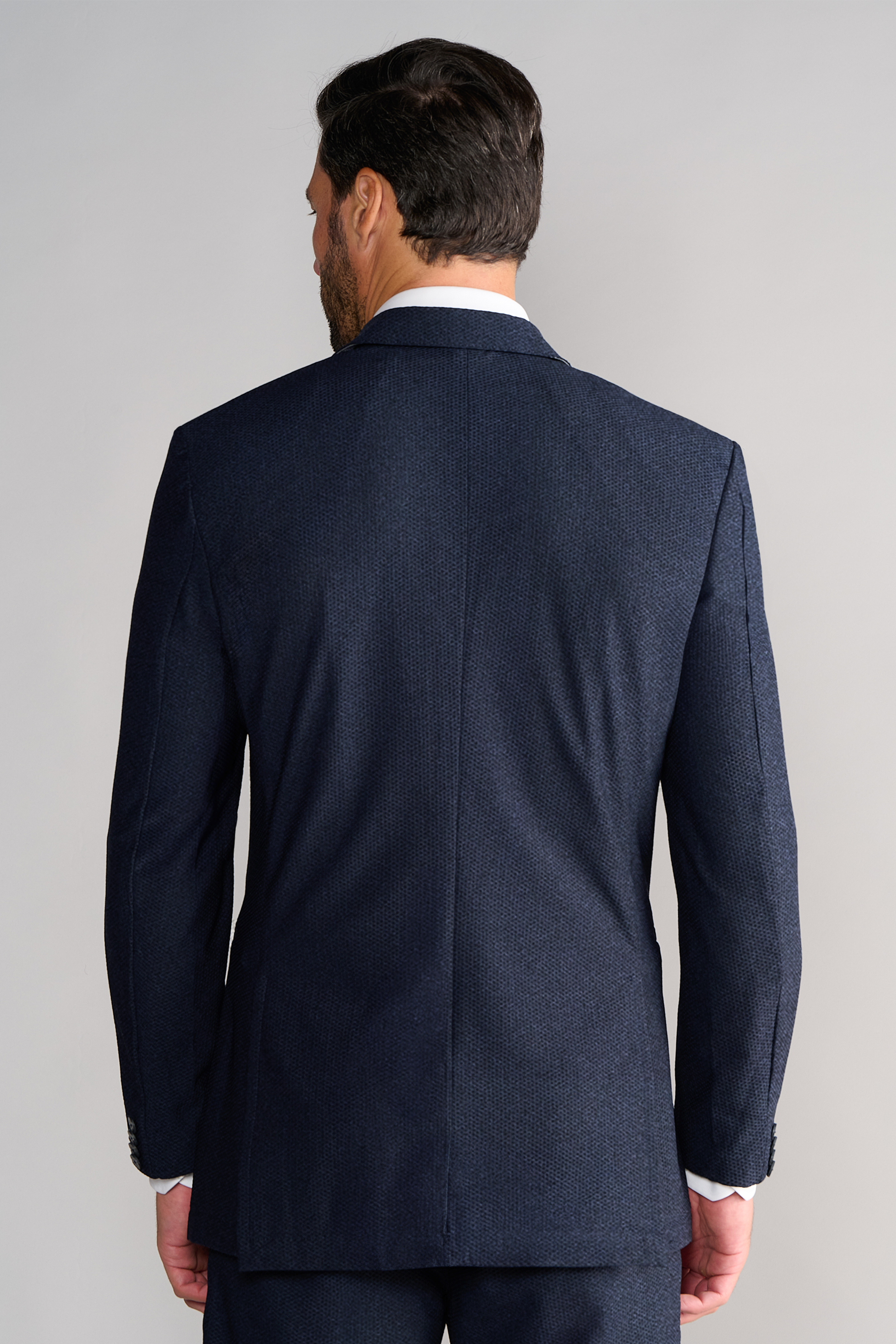 A man with short dark hair is seen from behind, wearing the Twillory AIR Blazer // Heather Navy. His hands rest by his sides, and the plain light gray background highlights the blazer’s formal texture and fit.