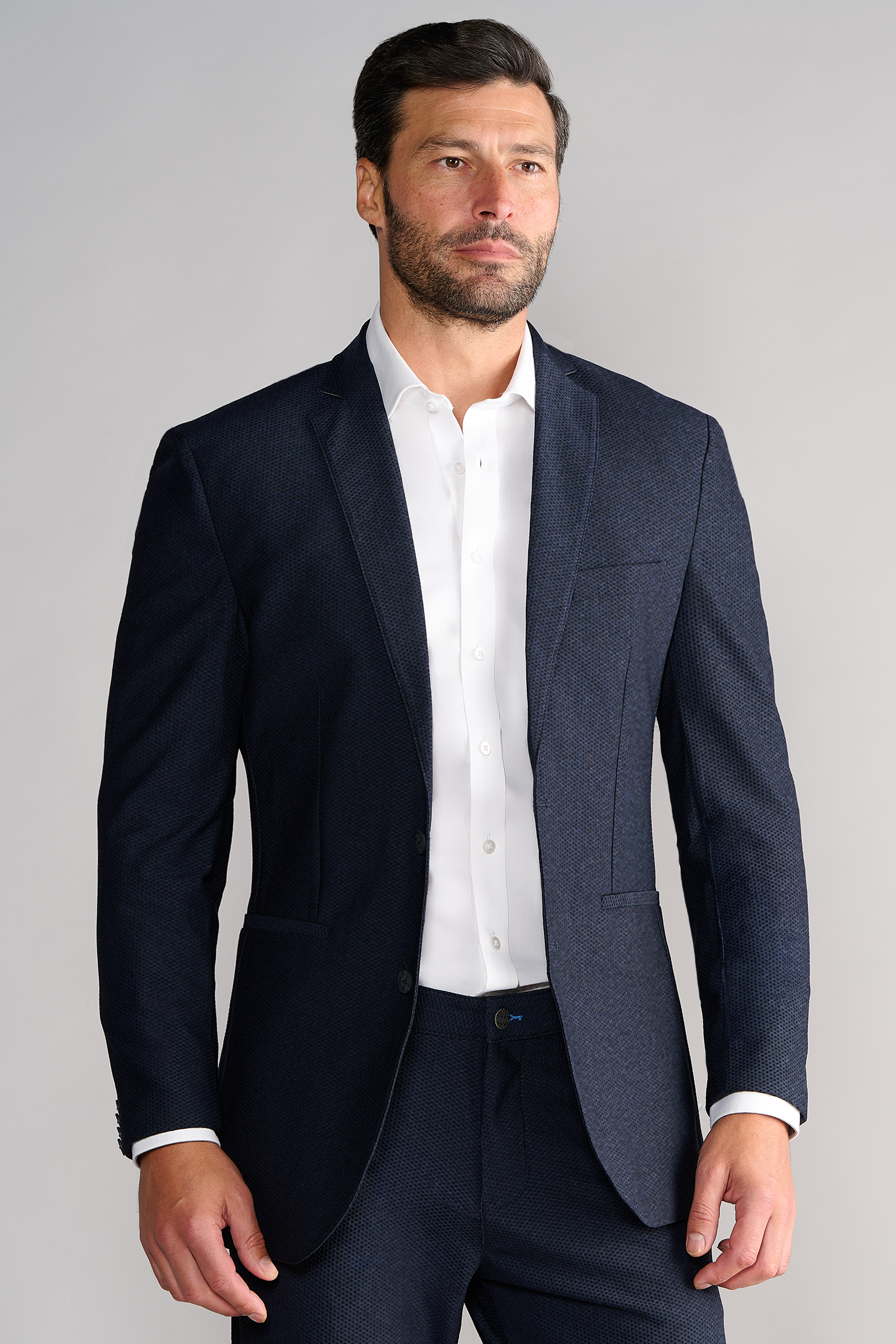 A man with short dark hair and a beard wears the Twillory AIR Blazer // Heather Navy, made from 360° Airflow Fabric, over a white dress shirt. He stands against a plain light gray background, looking slightly to the side.