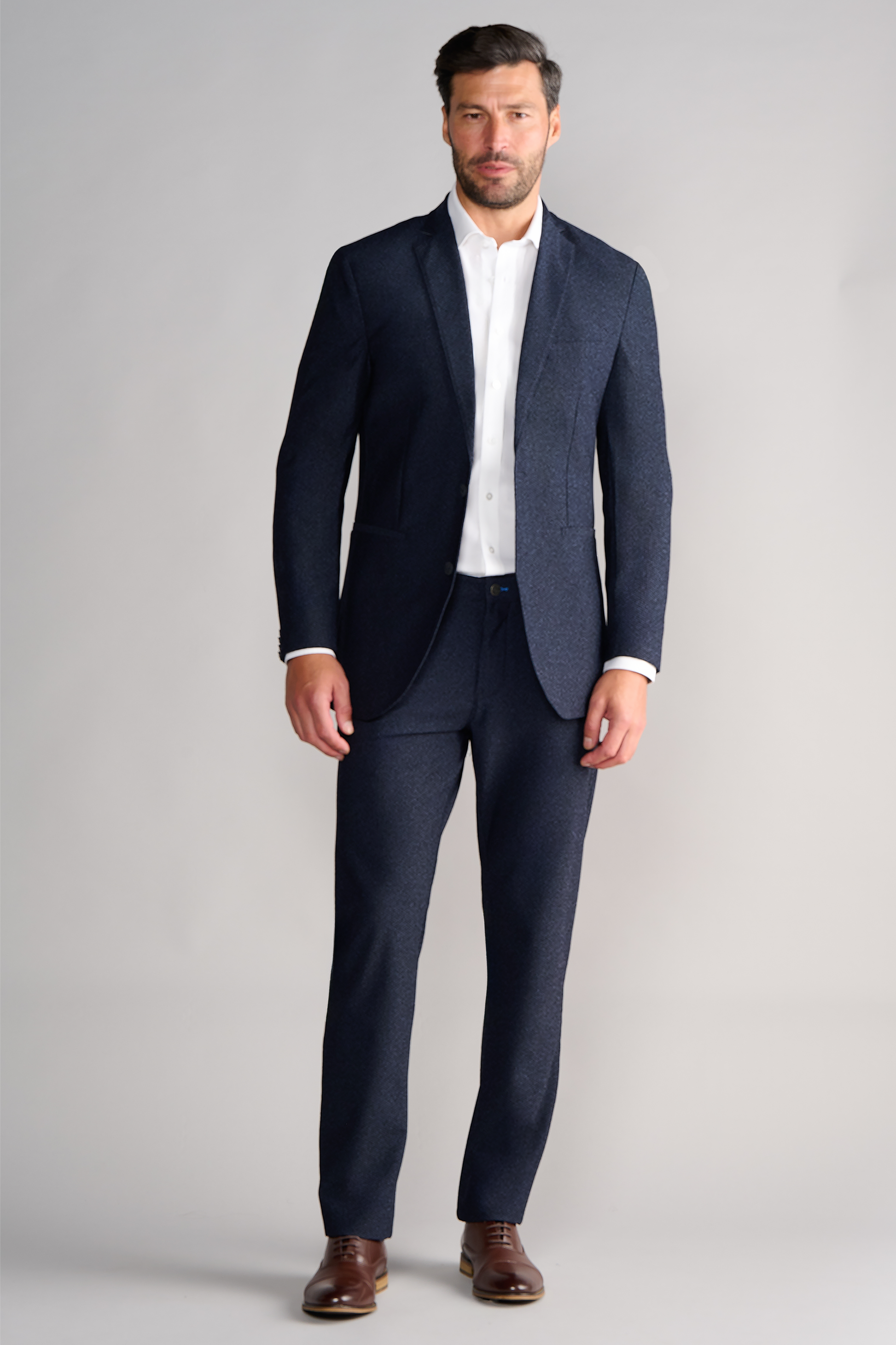 A man with short dark hair and a trimmed beard stands facing forward against a light gray background, wearing the Twillory AIR Blazer // Heather Navy, a white dress shirt, and brown leather dress shoes. His expression is neutral.