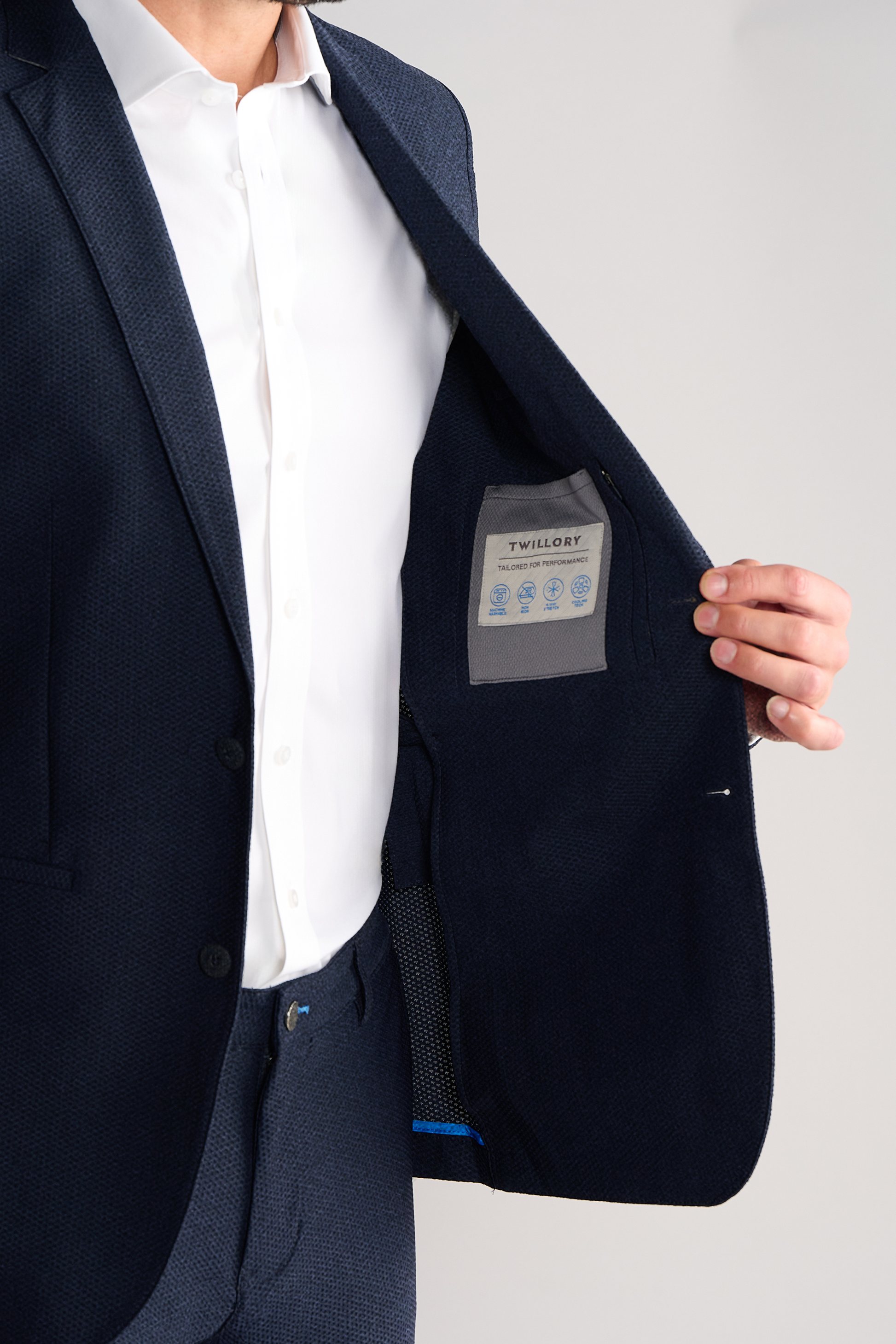 A man in a heather navy AIR Blazer by Twillory and a white shirt holds open his machine washable blazer, revealing the Twillory inner pocket tag with care instructions. The background is plain.