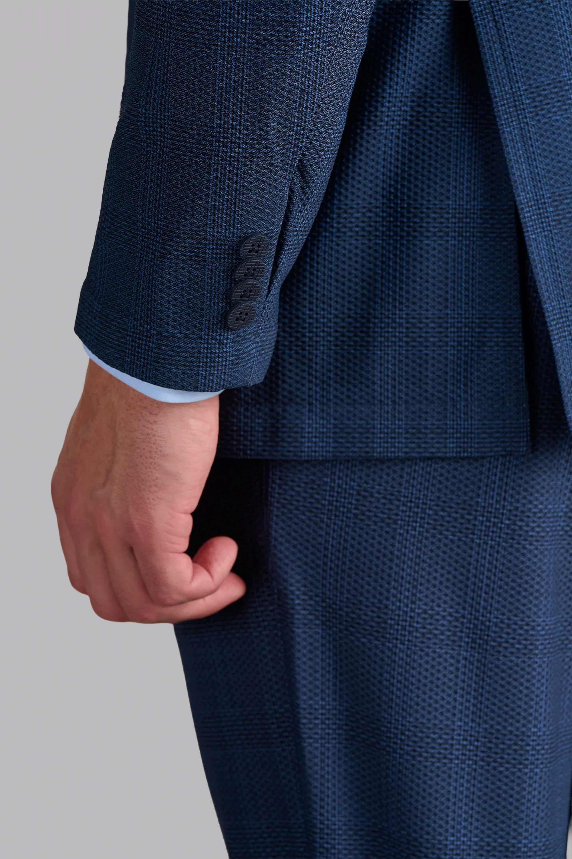 A close-up of a person’s left arm wearing the AIR Blazer // Blue Prince of Wales, featuring 360° Airflow Fabric with a subtle plaid pattern, three jacket sleeve buttons, and a light blue shirt cuff against a plain gray background.