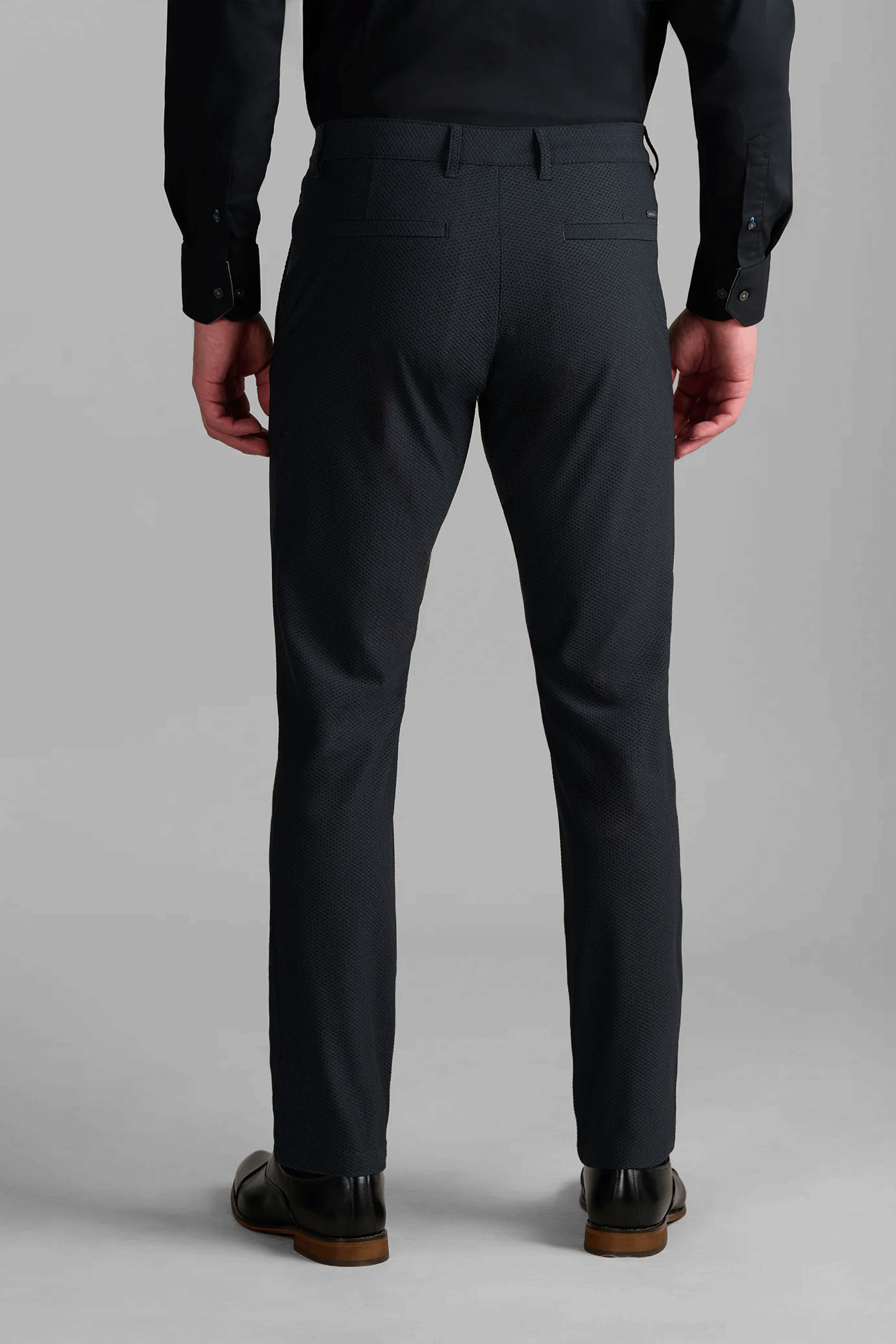 A person stands facing away, wearing Twillory AIR Pants in Heather Charcoal with two back welt pockets, paired with a black long-sleeve shirt and black dress shoes. The light gray background highlights their modern style.
