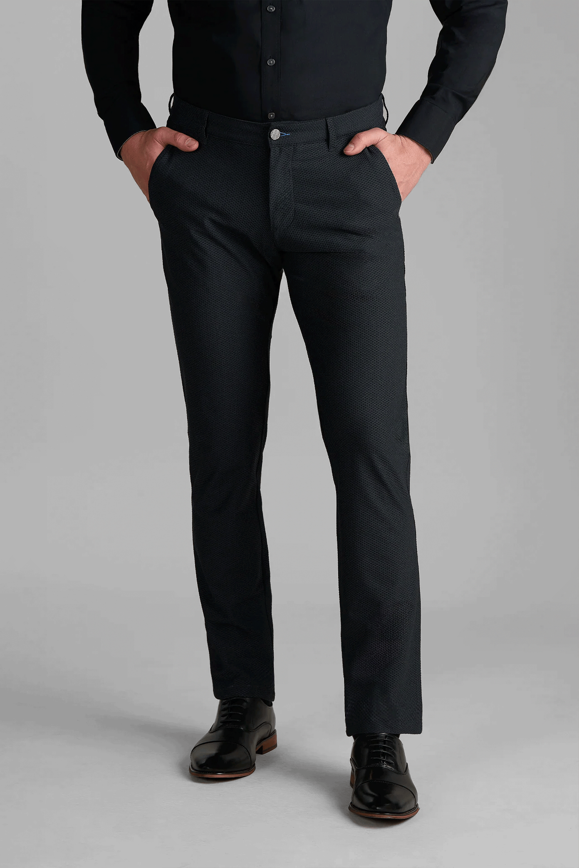 A person stands with hands in pockets, wearing Twillory AIR Pants in Heather Charcoal, a black button-up shirt, and shiny black dress shoes against a plain gray background. Only the body from shoulders down is visible.