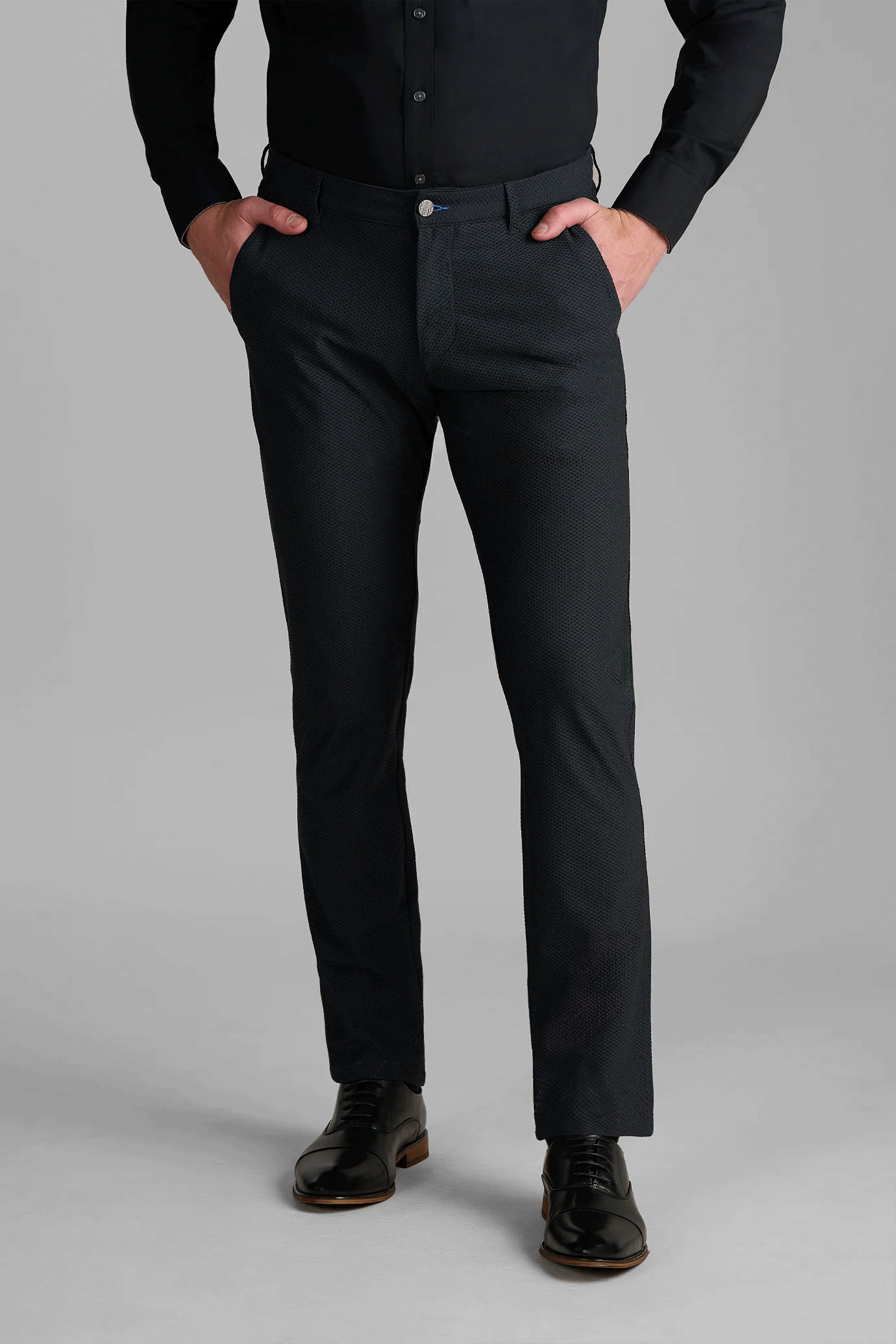 A person stands with hands in pockets, wearing Twillory AIR Pants in Heather Charcoal, a black button-up shirt, and shiny black dress shoes against a plain gray background. Only the body from shoulders down is visible.