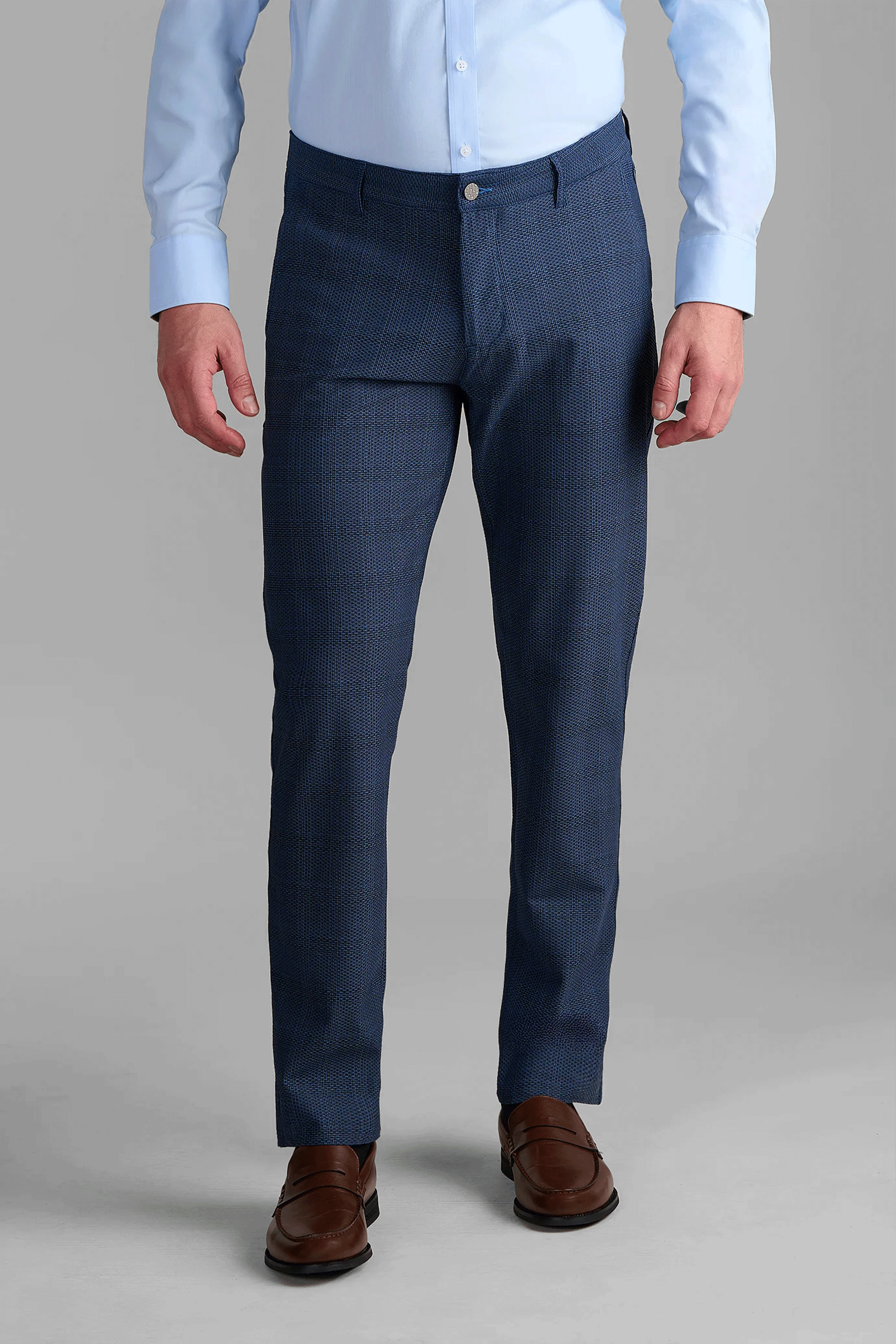 A man stands facing forward against a plain gray background, wearing a light blue dress shirt, AIR Pants // Blue Prince of Wales, and brown dress shoes.