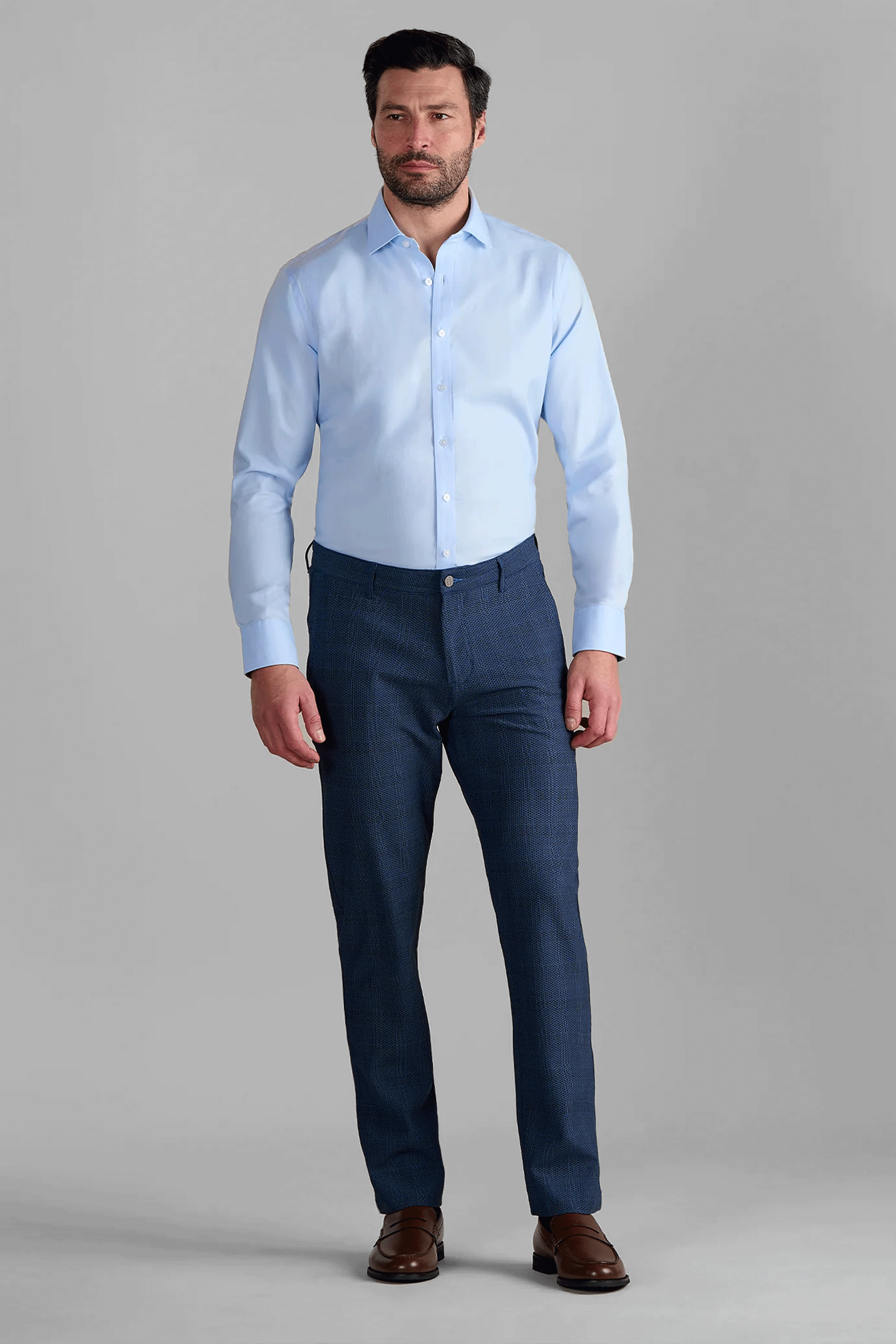 A man faces forward against a plain gray background, wearing a light blue dress shirt, AIR Pants // Blue Prince of Wales, and brown dress shoes.