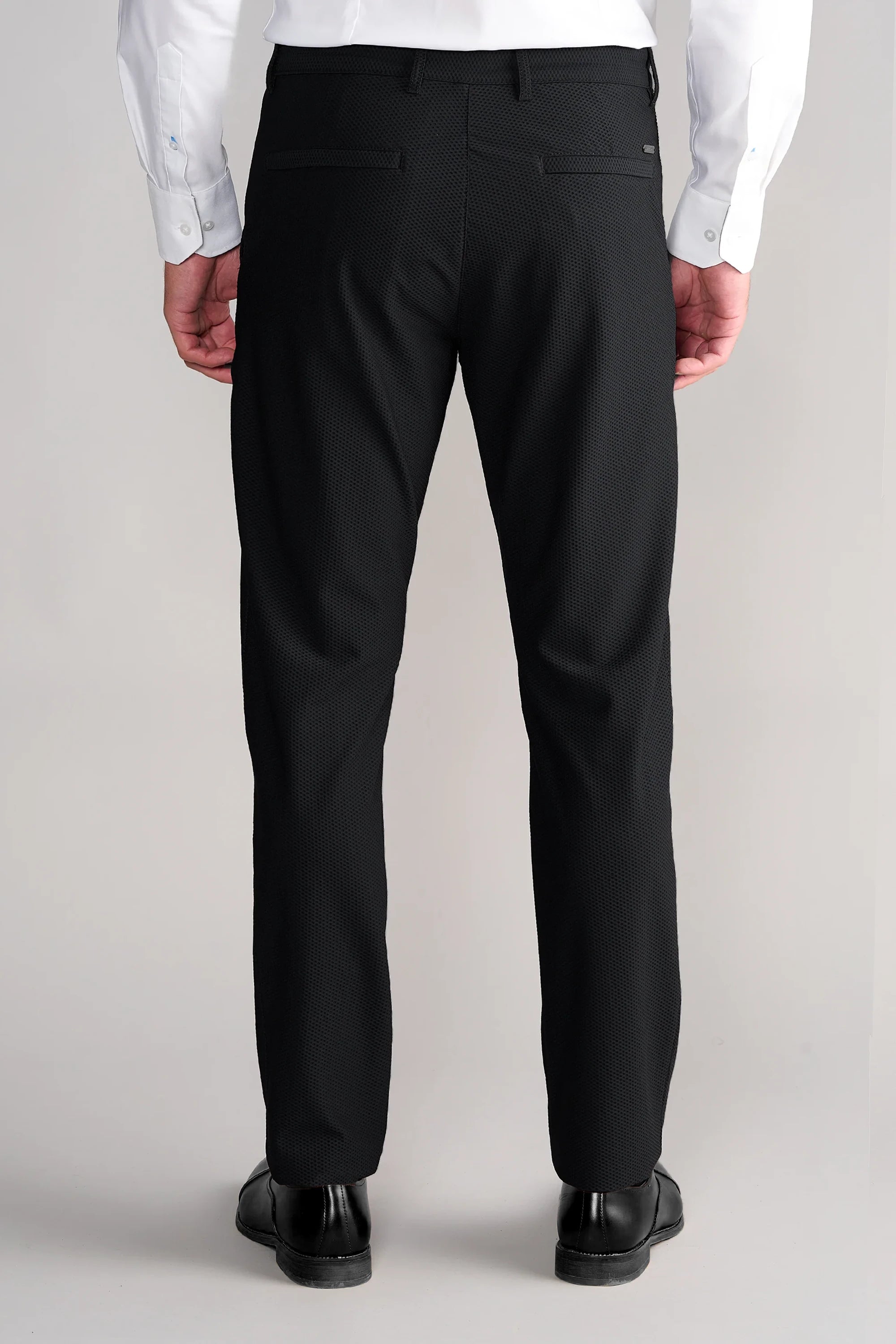 A person faces away from the camera, wearing a white dress shirt tucked into Twillory Gravity Pants // Black, featuring 4-way stretch, two back pockets, and belt loops. They also wear black dress shoes against a plain neutral background.