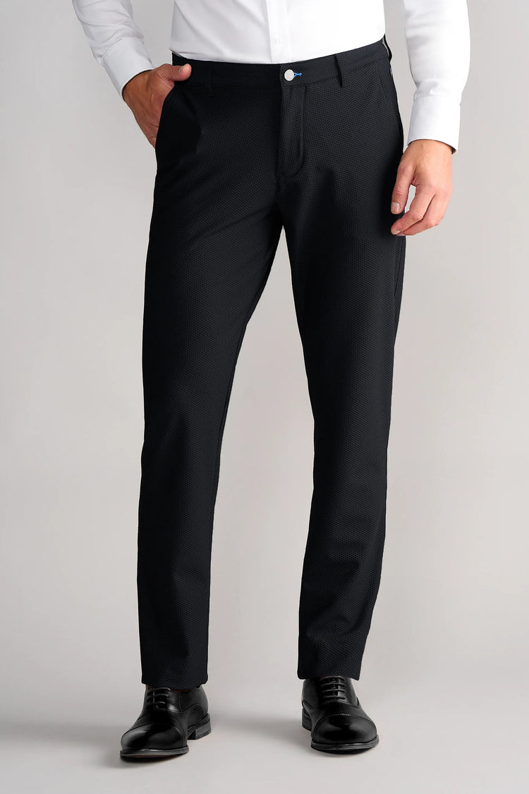 A man stands against a light gray background wearing Twillory Gravity Pants in black and black dress shoes, with a white long-sleeve shirt. His left hand is in his pocket; only his body from mid-torso to feet is visible.