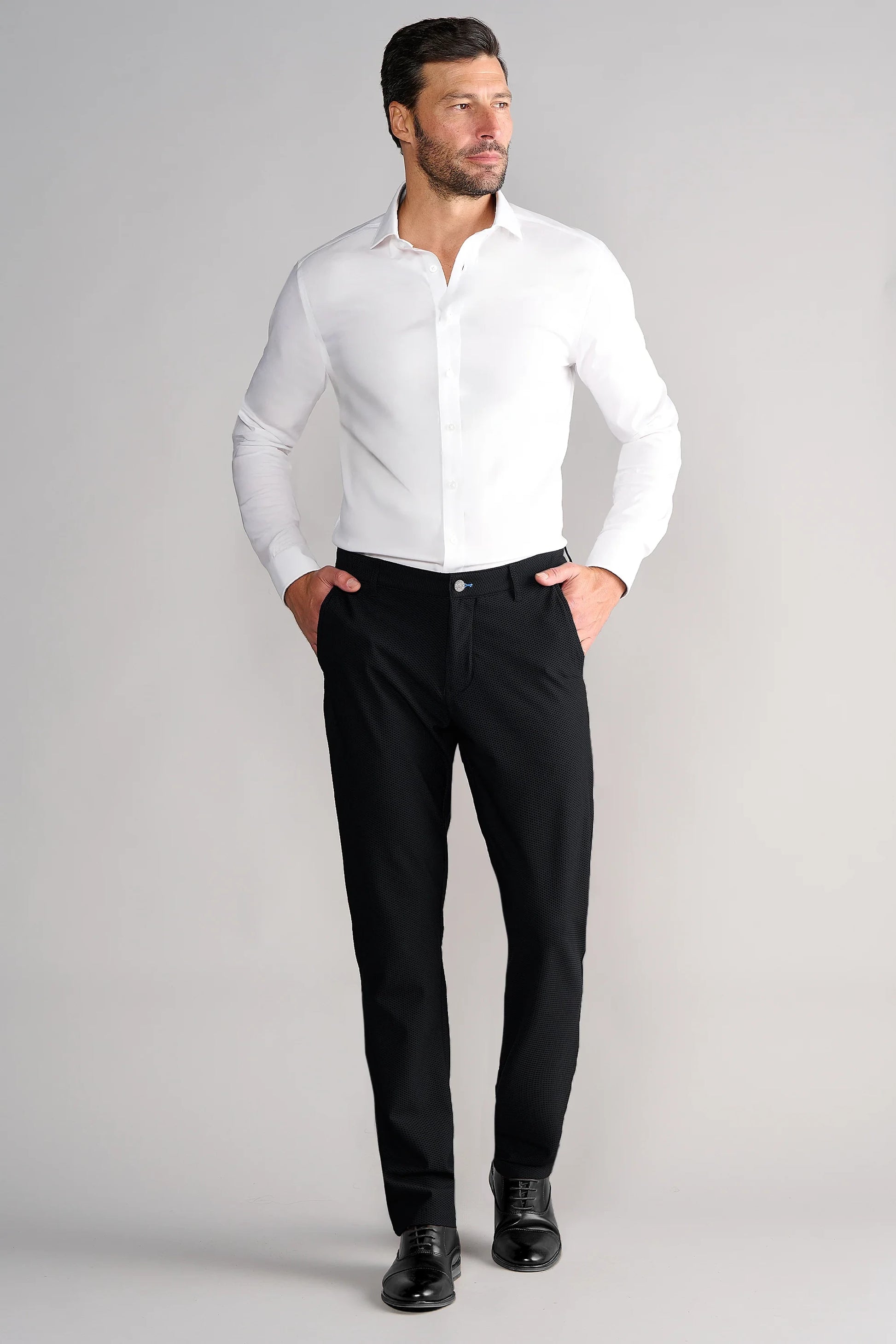 A man with short dark hair and a trimmed beard stands with hands in pockets, wearing a white dress shirt, Twillory Gravity Pants // Black, and black dress shoes. He looks slightly left against a plain light gray background.