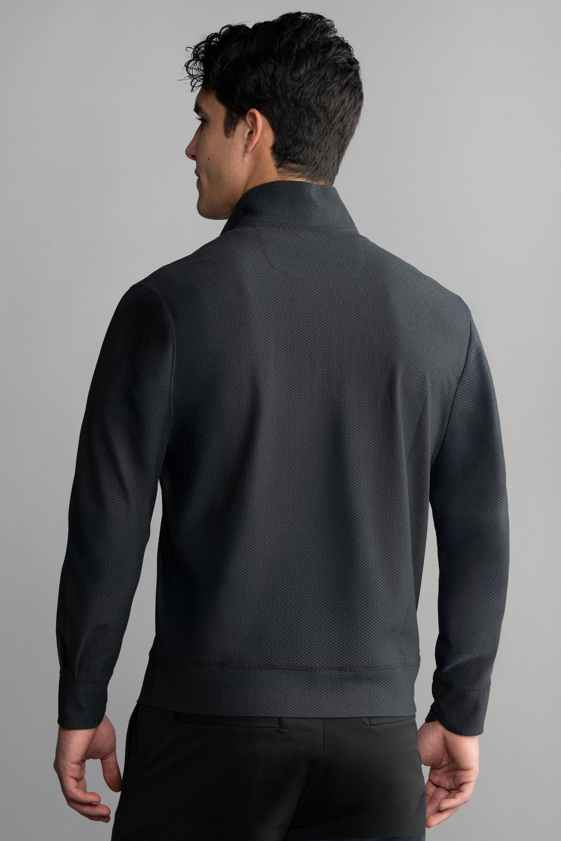 A man with short dark hair is seen from the back wearing the Twillory Air Quarter Zip in Charcoal, featuring a textured finish, high collar, and long sleeves. He stands against a plain gray background with his left hand by his side.