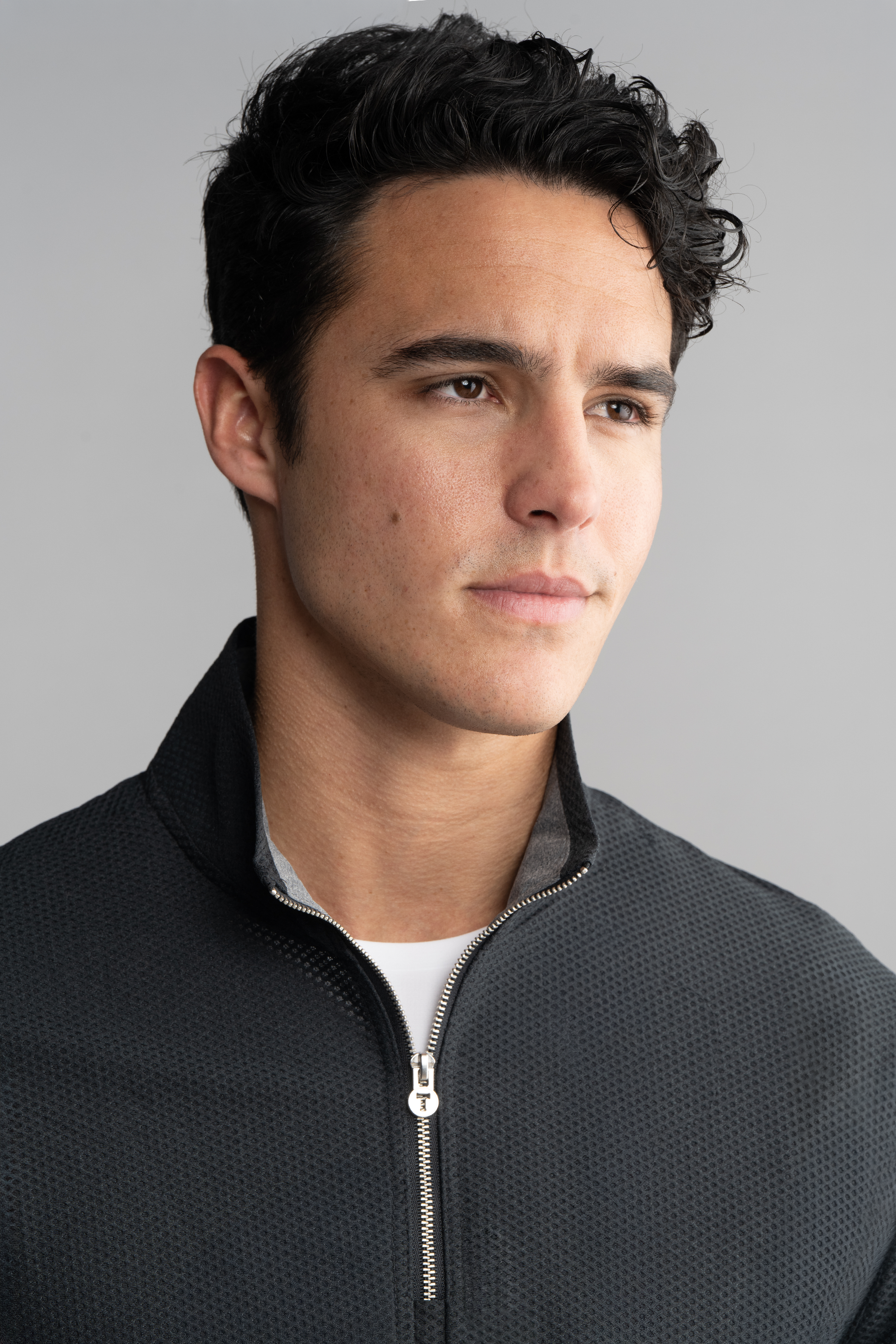 A young man with short dark hair and a light complexion looks slightly right, wearing the Twillory Air Quarter Zip in Charcoal over a white shirt. The background is plain light gray with soft, even lighting.