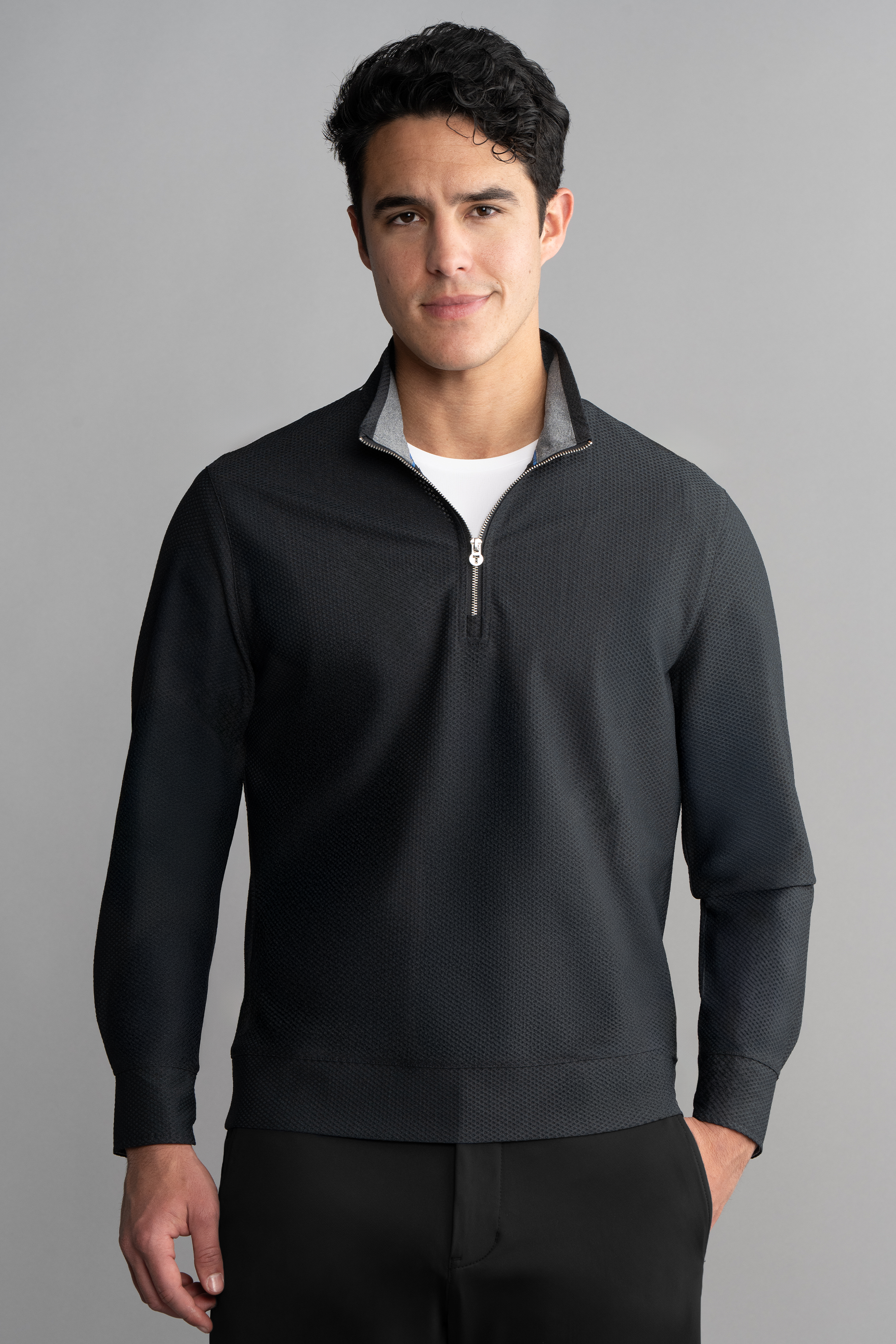 A man with short, curly dark hair stands against a plain gray background, wearing the Twillory Air Quarter Zip in Charcoal over a white T-shirt. He smiles slightly with his hands relaxed at his sides.