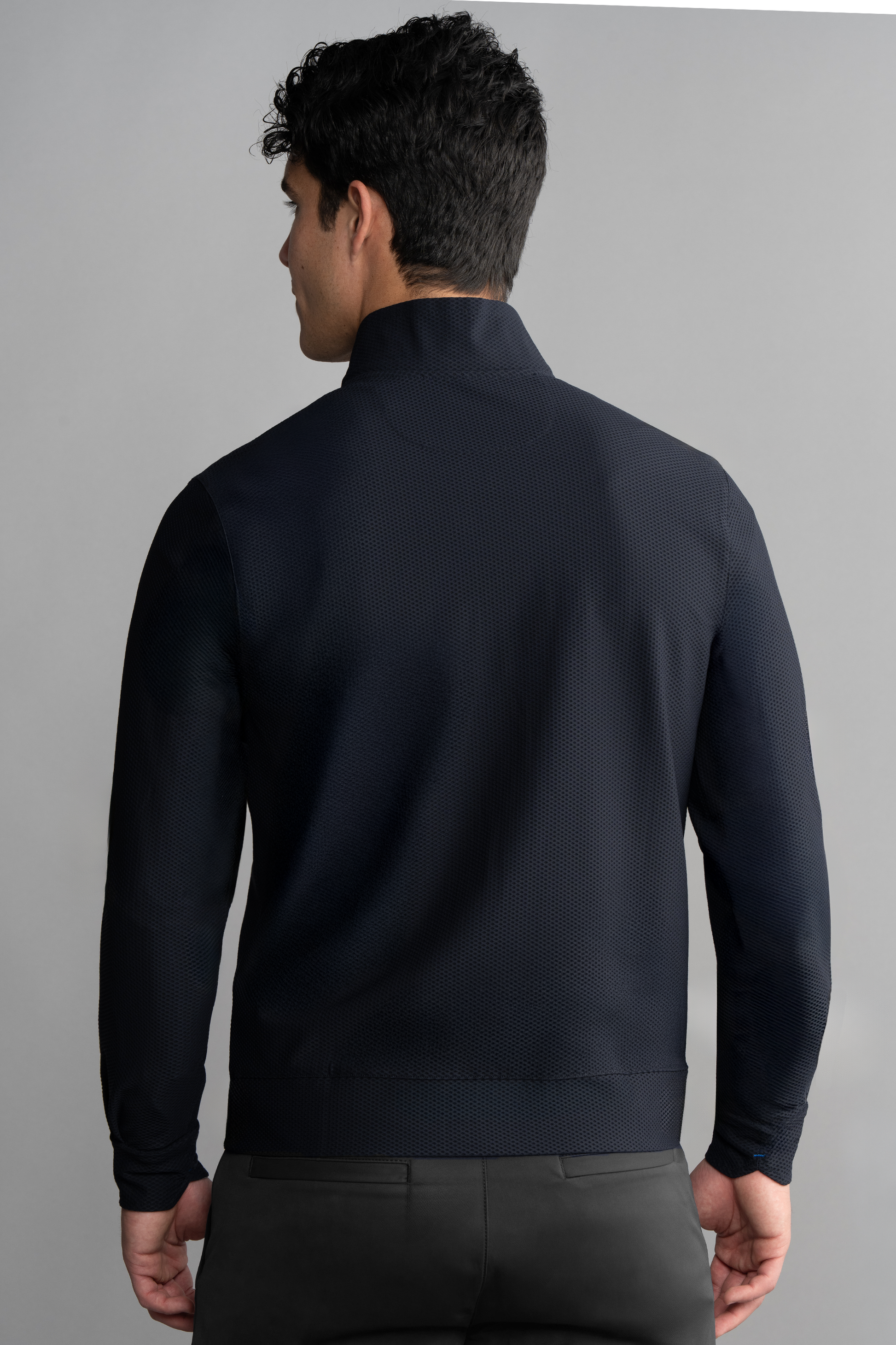 A man with short dark hair, seen from behind, wears the Twillory Air Quarter Zip in navy with a stand-up collar and black pants. His left arm is slightly bent as he stands against a plain light gray background.