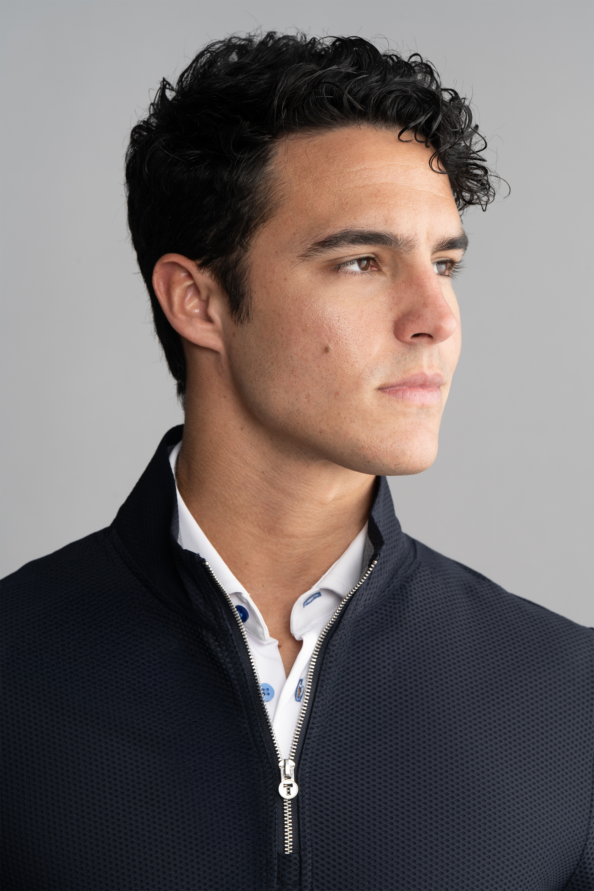 A young man with short, curly dark hair looks right, wearing the Twillory Air Quarter Zip in Navy—crafted from wrinkle-free AIR fabric—over a white collared shirt with blue accents, set against a plain light gray background.