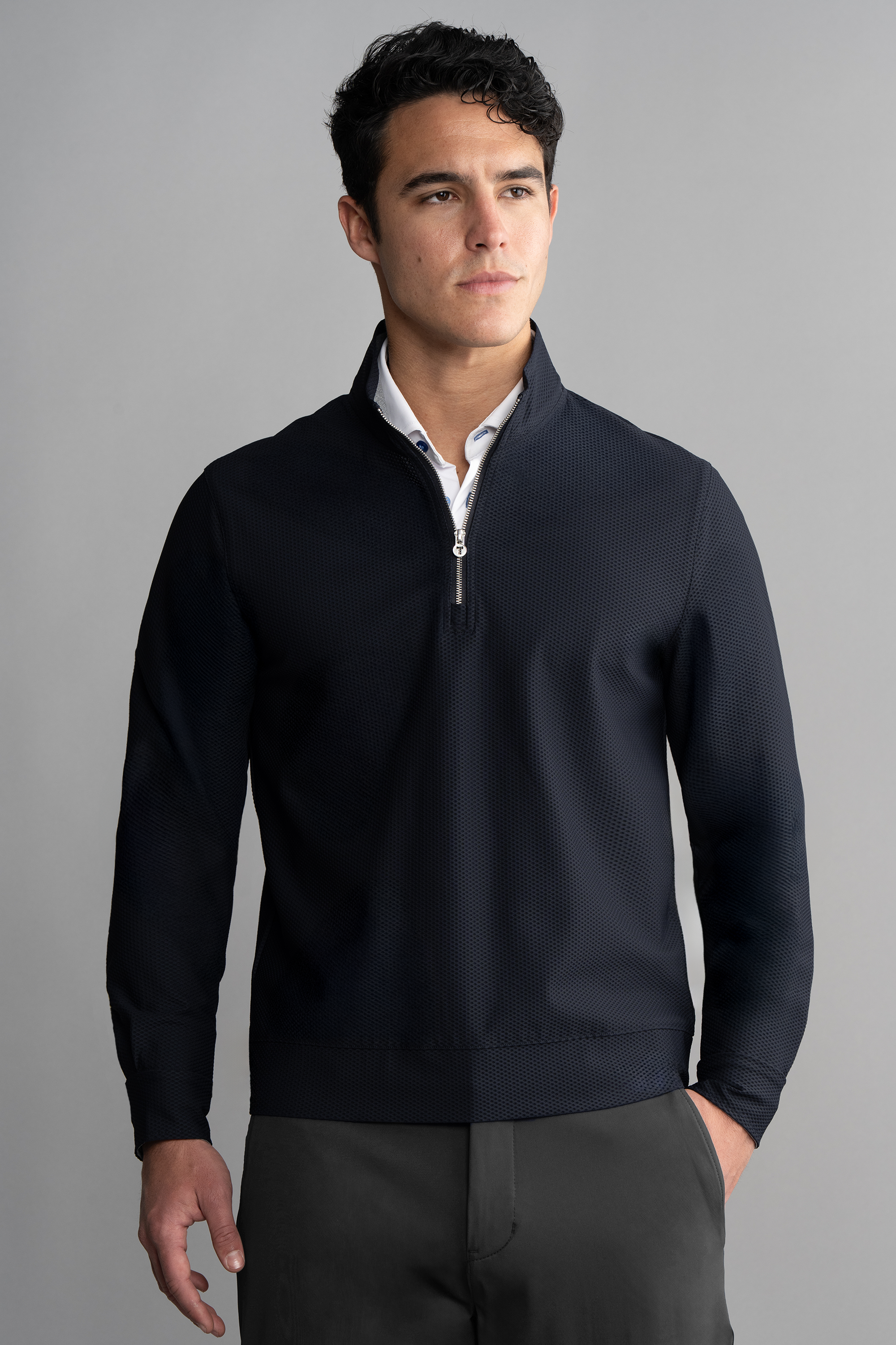 A young man with short dark hair wears the Twillory Air Quarter Zip in Navy over a white collared shirt and dark pants. He stands against a gray background, looking slightly to the side with a neutral expression, one hand in his pocket.