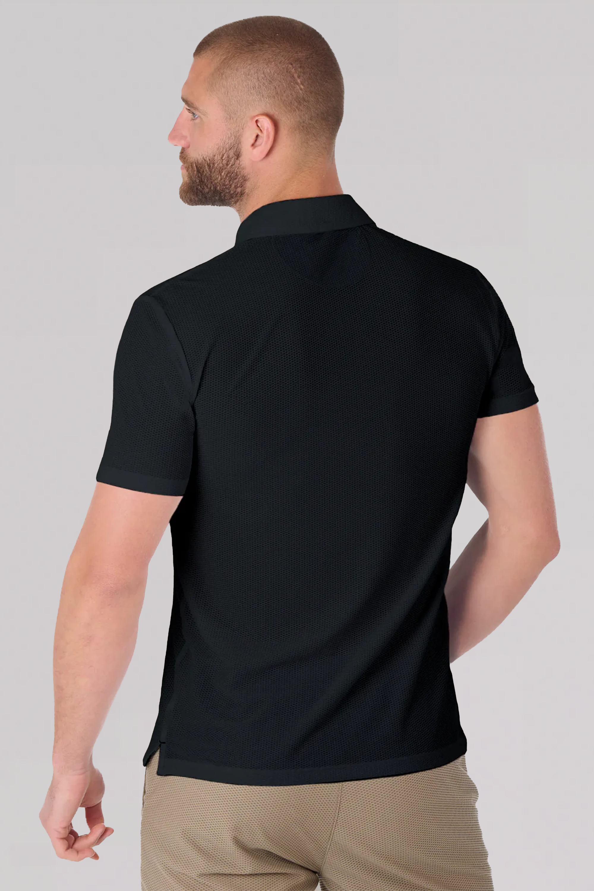 A man with short hair and a beard is seen from behind, wearing the Twillory AIR Zip Polo Solid in black and light brown pants. He stands against a plain, light background, his head slightly turned to the side.