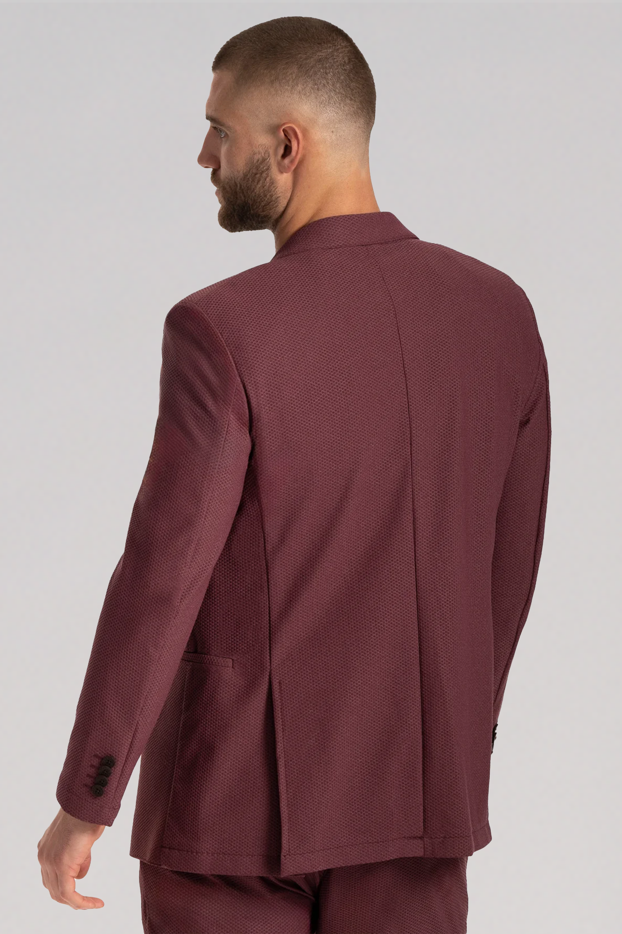 A man with short hair and a beard is seen from behind in Twillory’s AIR Blazer // Burgundy, made with 360° Airflow Fabric, and matching trousers. He faces left, arm slightly bent, against a plain light gray background.