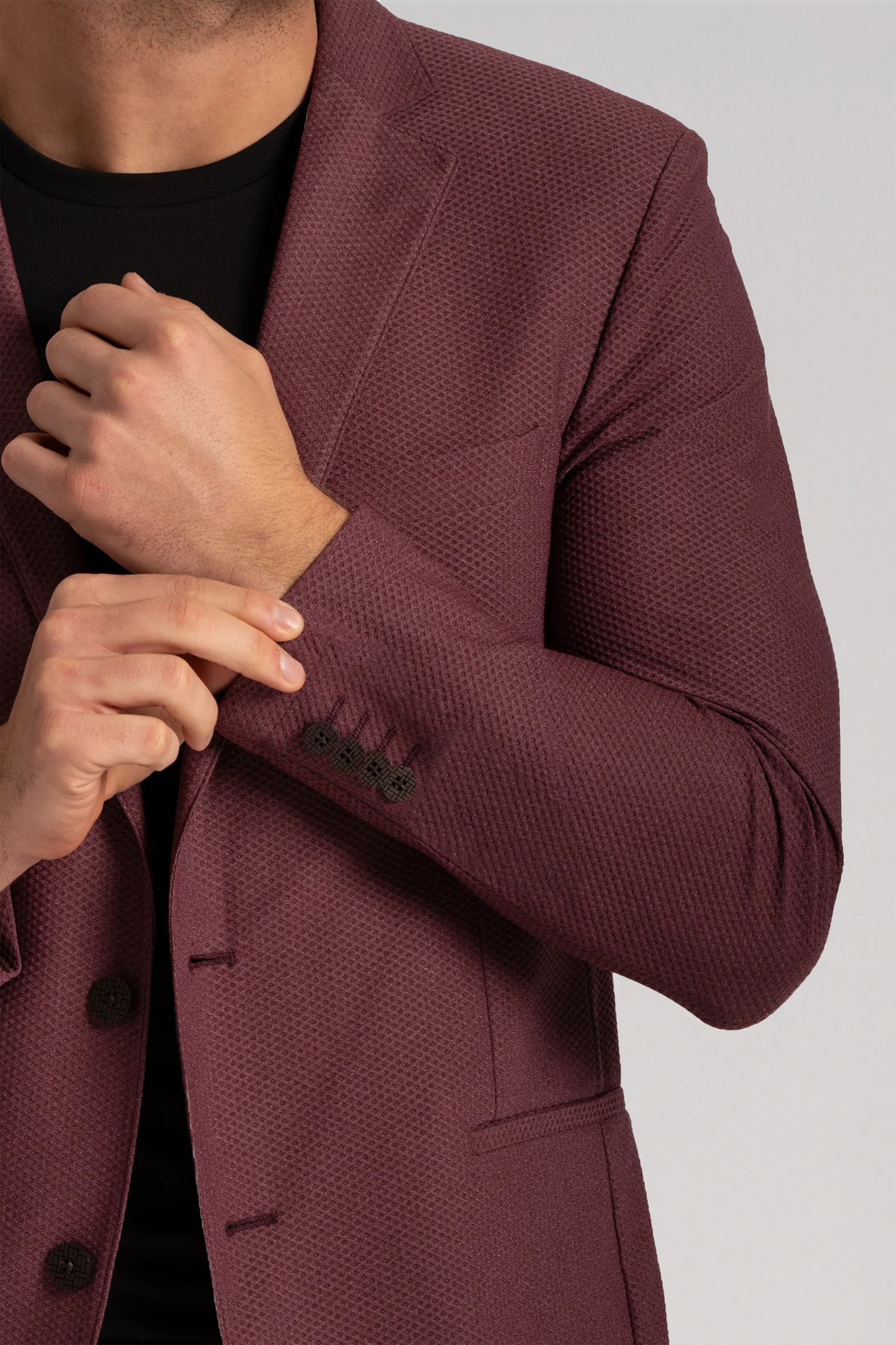 A person adjusts the sleeve cuff of the Twillory AIR Blazer // Burgundy, made from performance fabric, over a black shirt. The cropped image focuses on the upper torso and arms, highlighting black buttons on the cuff and front.