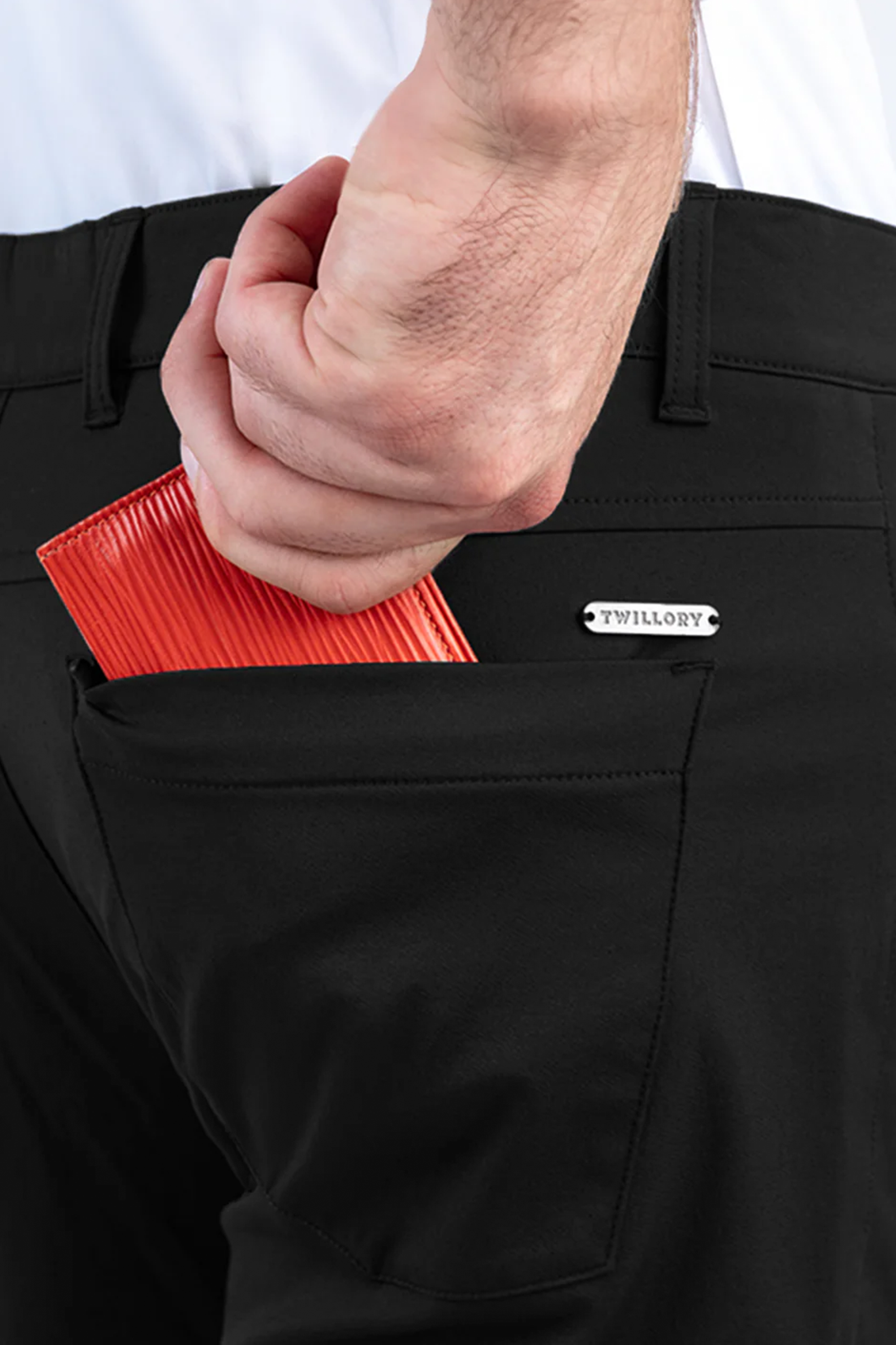A person places a red wallet into the back pocket of Performance Five Pocket // BLACK pants with a visible Twillory label, while wearing a tucked-in white, sweat-wicking shirt.