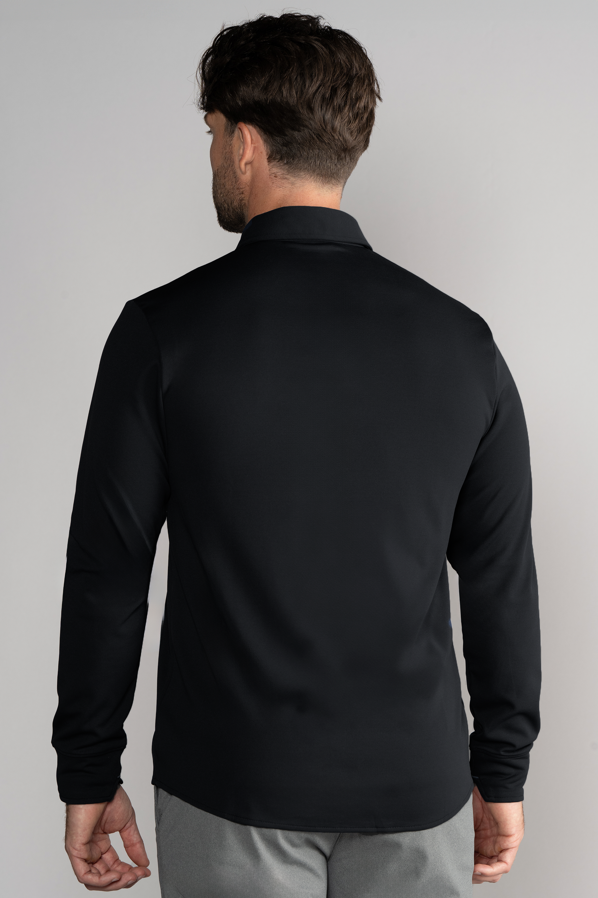 A man with short brown hair is seen from the back wearing the Twillory Long Sleeve Button Down Polo Solid in black and light gray pants, standing against a plain light gray background with his arms relaxed at his sides.
