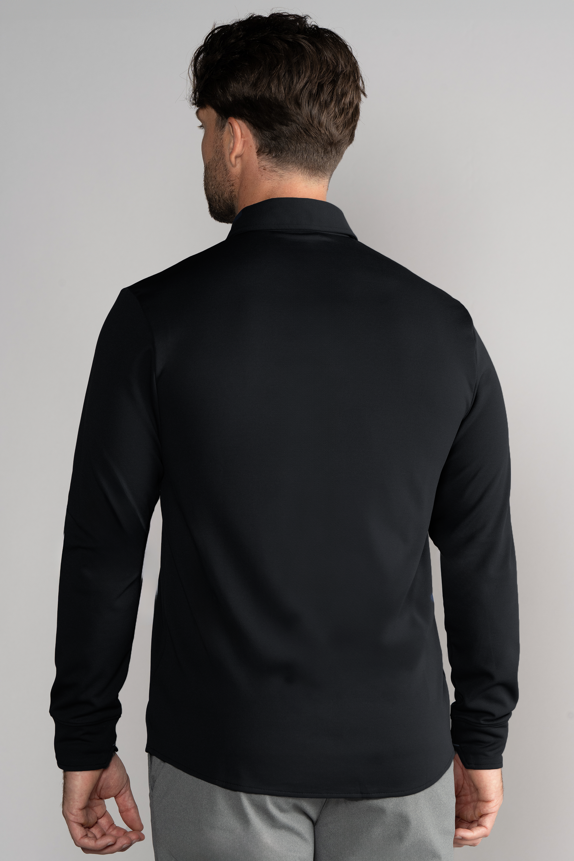 A man with short brown hair is seen from the back wearing the Twillory Long Sleeve Button Down Polo Solid in black and light gray pants, standing against a plain light gray background with his arms relaxed at his sides.