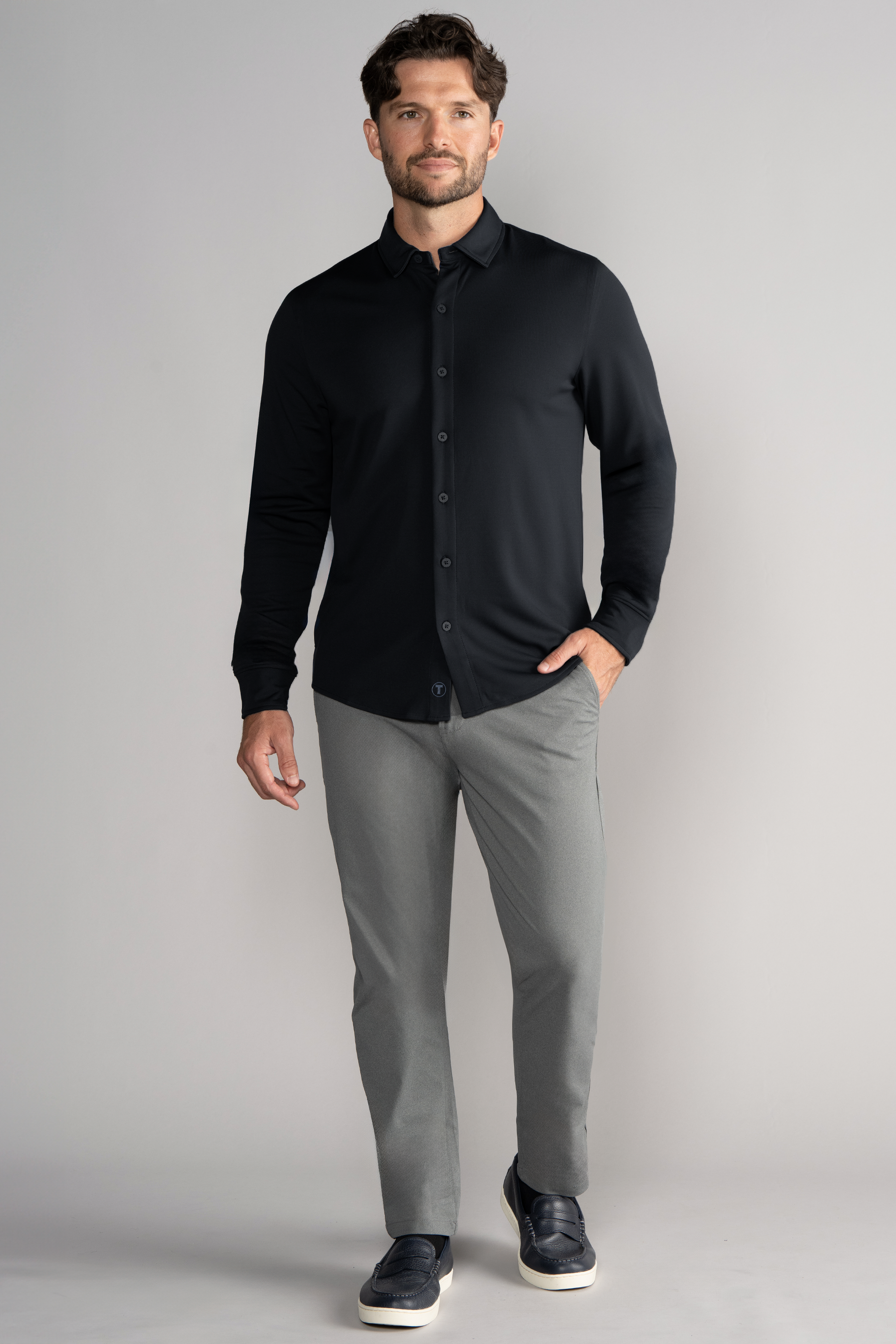 A man with short brown hair and a beard stands against a plain light gray background, smiling slightly with one hand in his pocket. He wears the Twillory Long Sleeve Button Down Polo Solid in black, light gray slim-fit pants, and dark slip-on shoes.