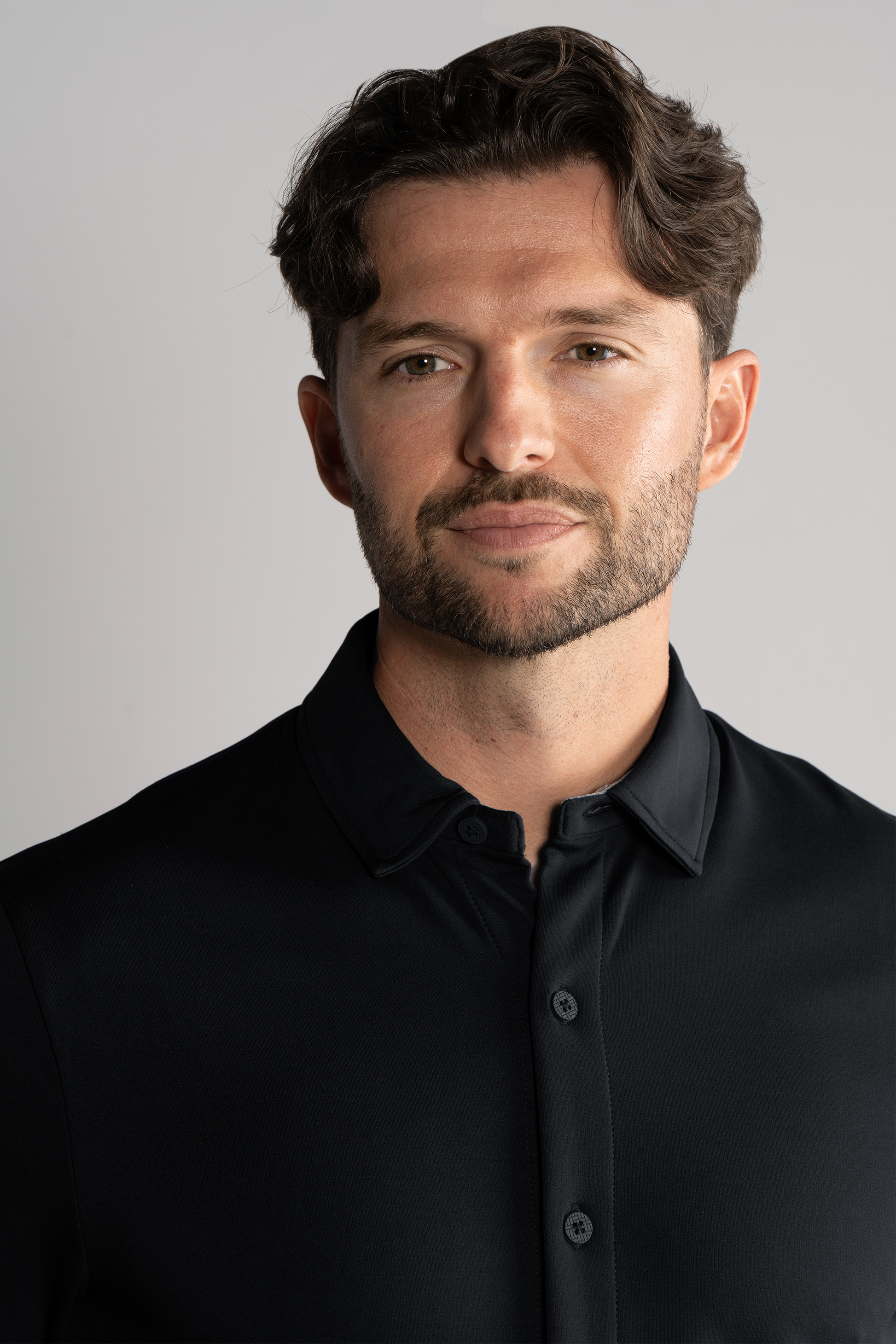 A man with wavy brown hair and a trimmed beard wears the Twillory Long Sleeve Button Down Polo Solid in black. He faces forward and smiles slightly, set against a neutral gray background with soft lighting highlighting his features.