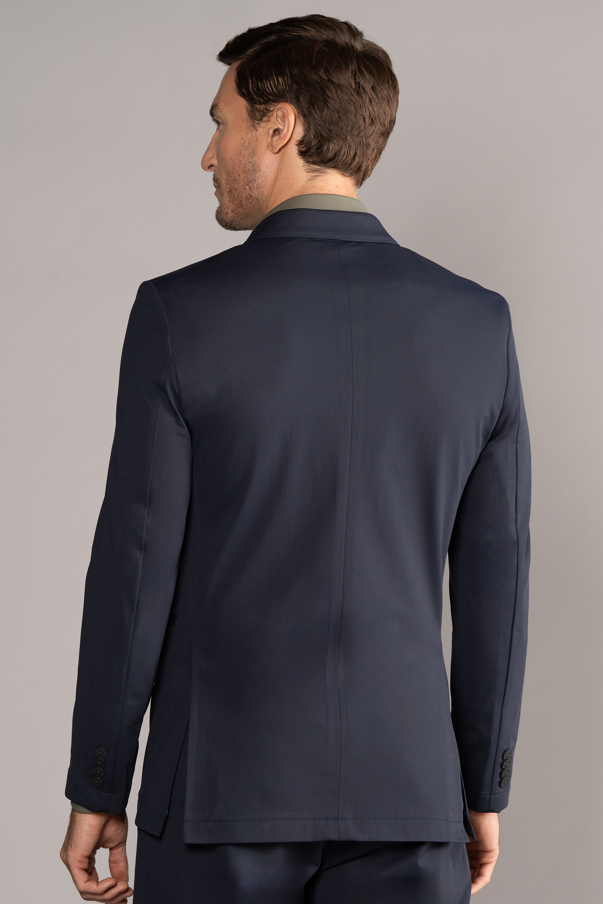 A man with short brown hair models the Twillory Performance Double Breasted Blazer in navy, paired with matching pants, standing mid-thigh up against a plain light gray background with his left arm slightly bent at his side.