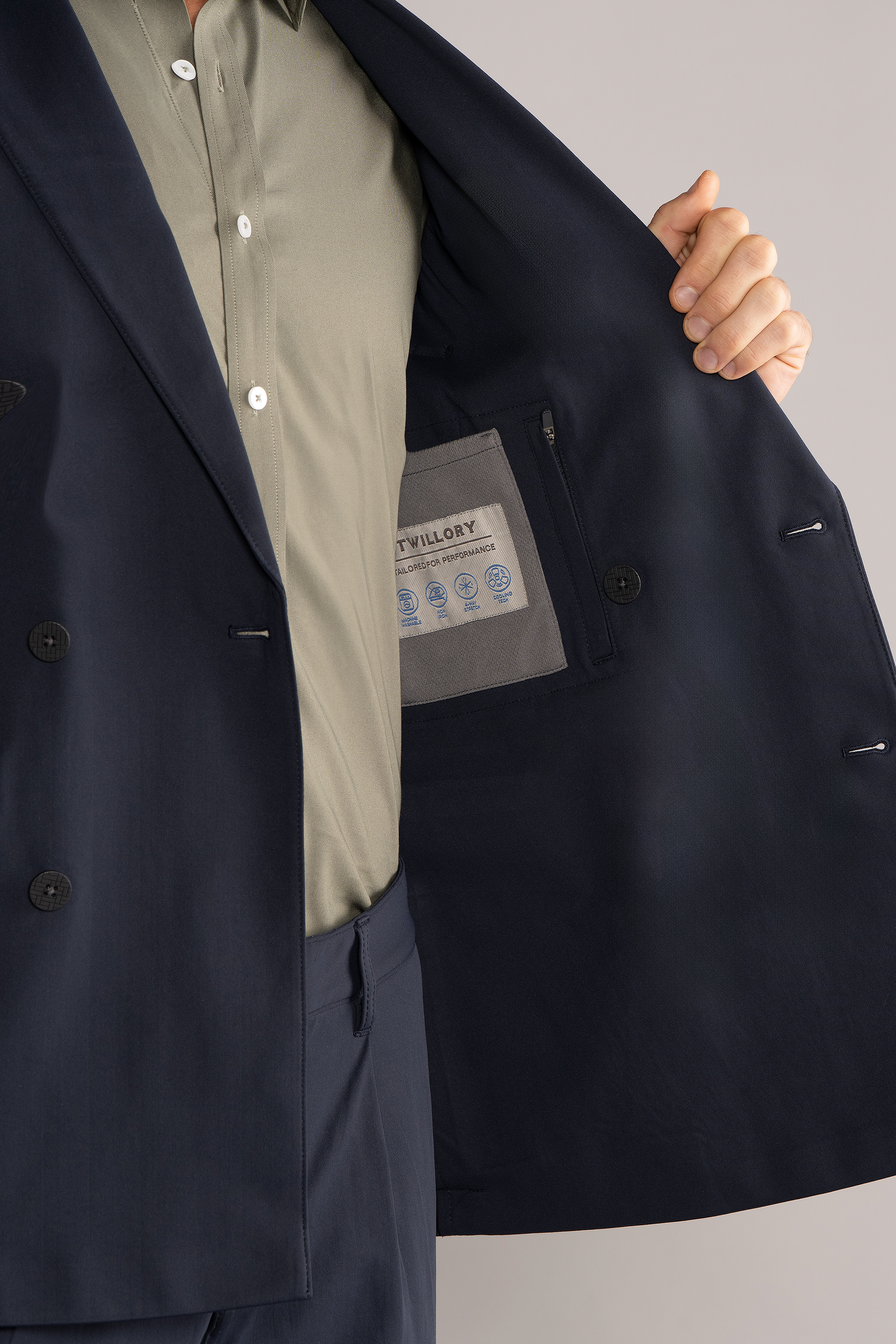 A person wears the Twillory Performance Double Breasted Blazer // Navy, made from Japanese stretch fabric, with a light green shirt. They hold open the machine washable blazer to show the care label. Face is not visible; plain light background.