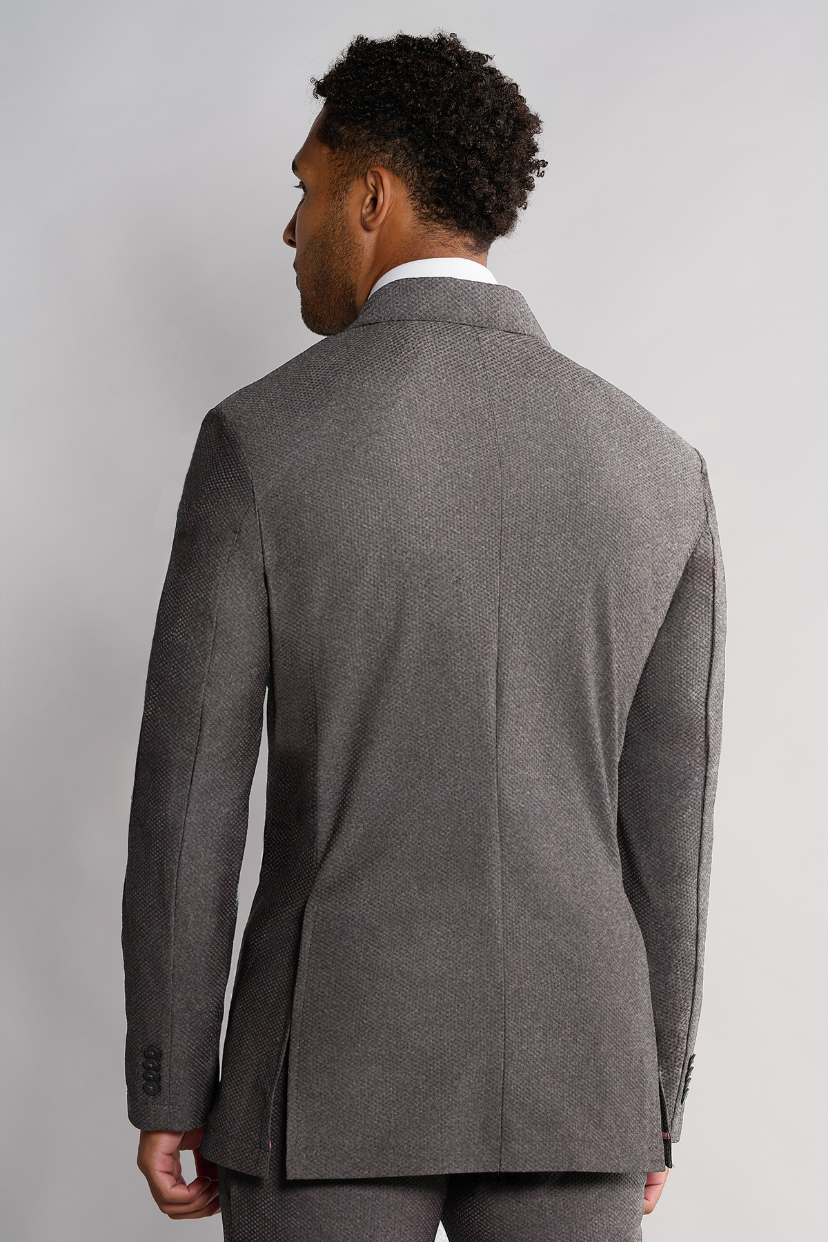 A man with short curly hair, seen from behind, wears the Twillory AIR Blazer // Heather Light Grey. This tailored-fit, machine-washable blazer features 360° Airflow Fabric, a notch lapel, and sleeve buttons. Background is plain light gray.