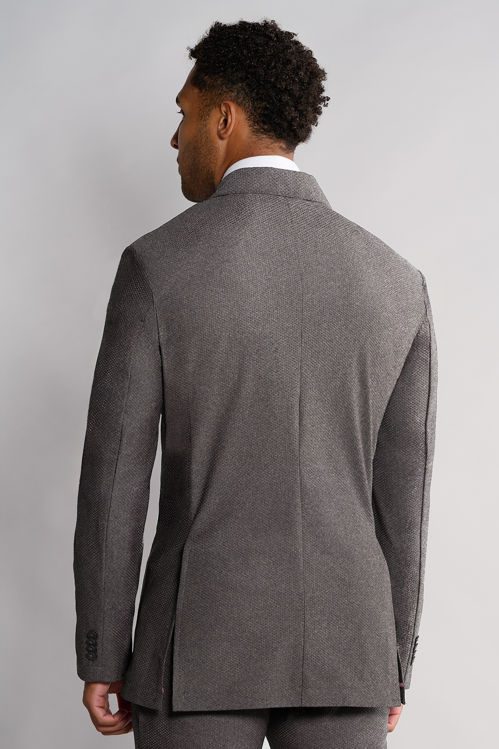 A man with short curly hair, seen from behind, wears the Twillory AIR Blazer // Heather Light Grey. This tailored-fit, machine-washable blazer features 360° Airflow Fabric, a notch lapel, and sleeve buttons. Background is plain light gray.