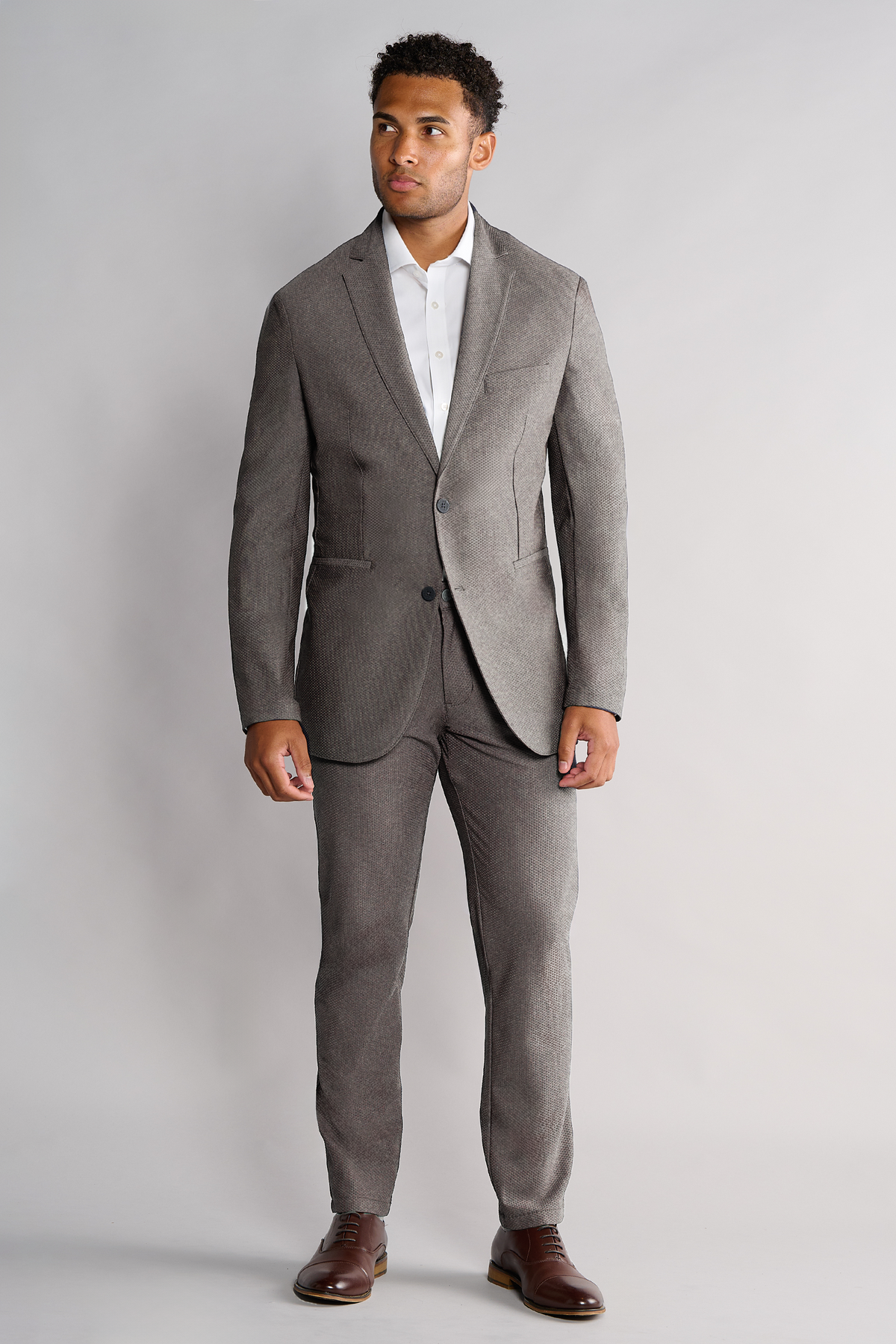 A man stands facing forward, wearing Twillory's AIR Blazer in Heather Light Grey with matching trousers and a white dress shirt. His hands are relaxed at his sides as he looks slightly to his left, set against a plain light background.