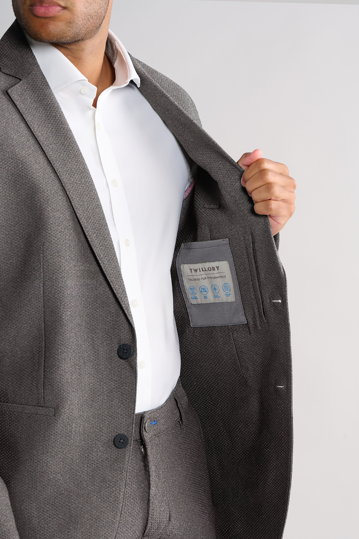 A man in a gray AIR Blazer // Heather Light Grey by Twillory opens his jacket to display the brand label, showcasing the luxe 360° Airflow Fabric and easy-care design. Only his lower face is visible.