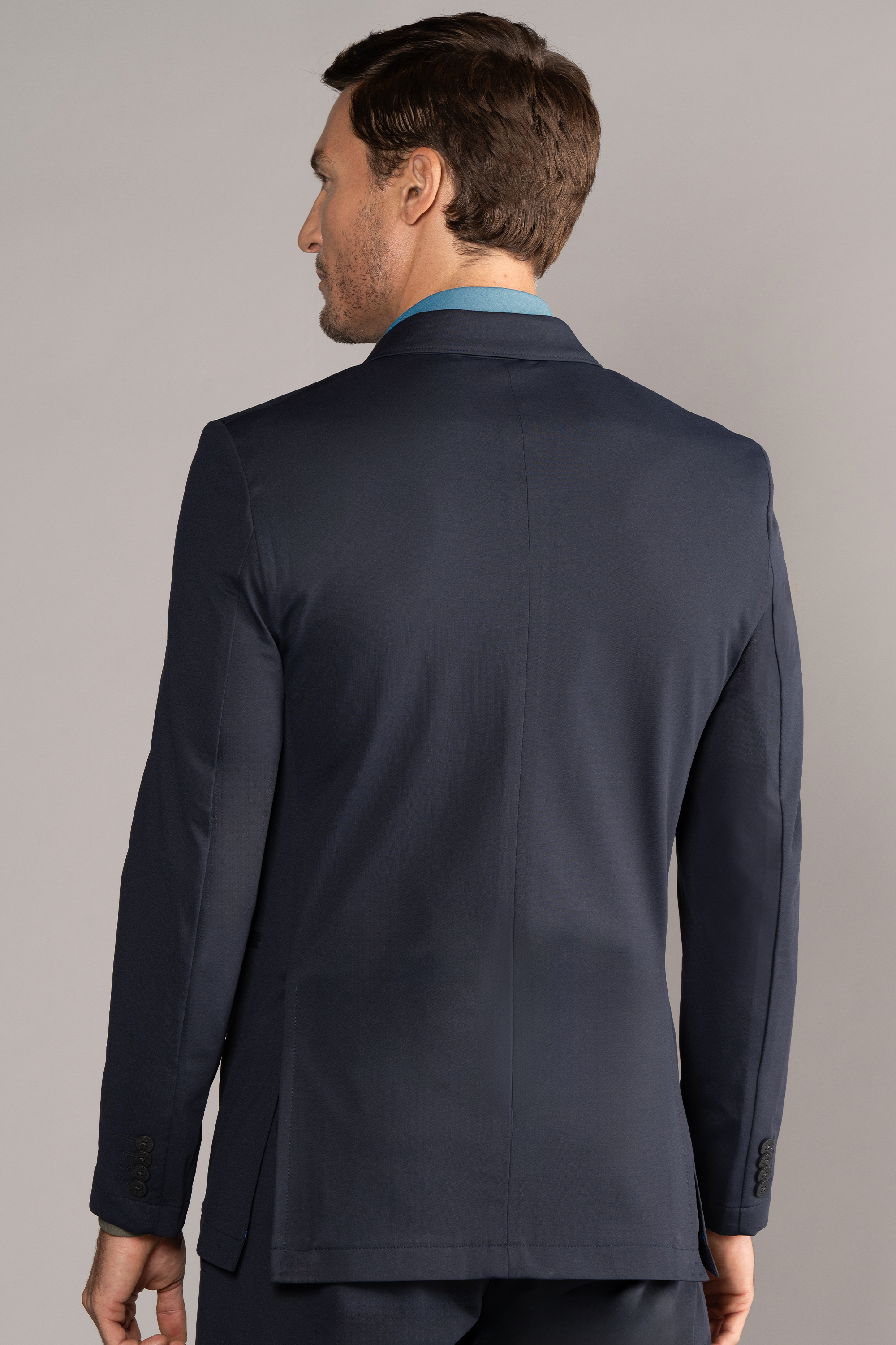A man with short brown hair is seen from behind wearing Twillory’s Performance Patch Pocket Blazer // Navy, paired with matching pants. The plain background accentuates the blazer’s fit, clean lines, seam details, and buttoned cuffs.