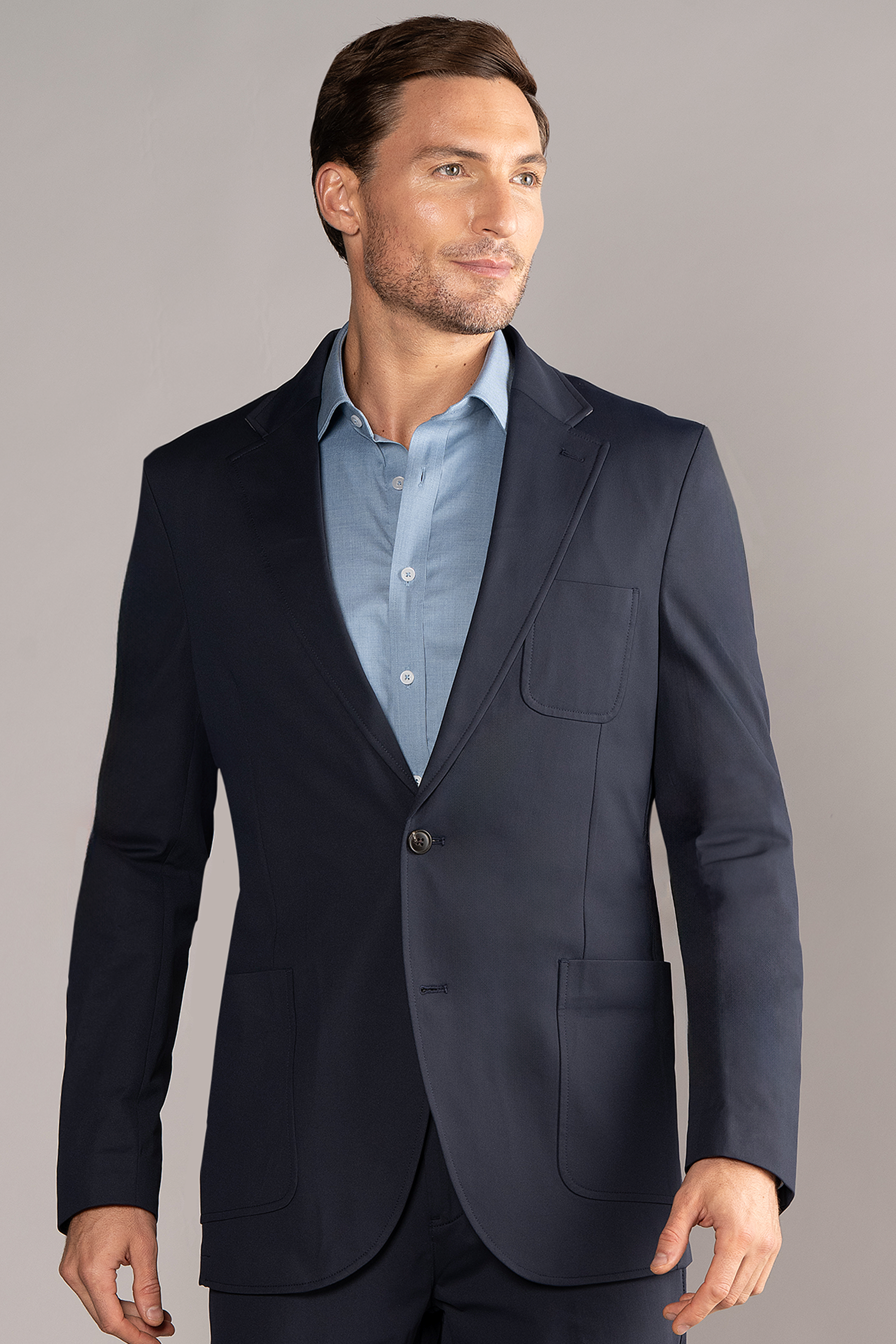 A man with short brown hair and light stubble wears the Twillory Performance Patch Pocket Blazer in navy over a light blue shirt, standing confidently against a light gray background.