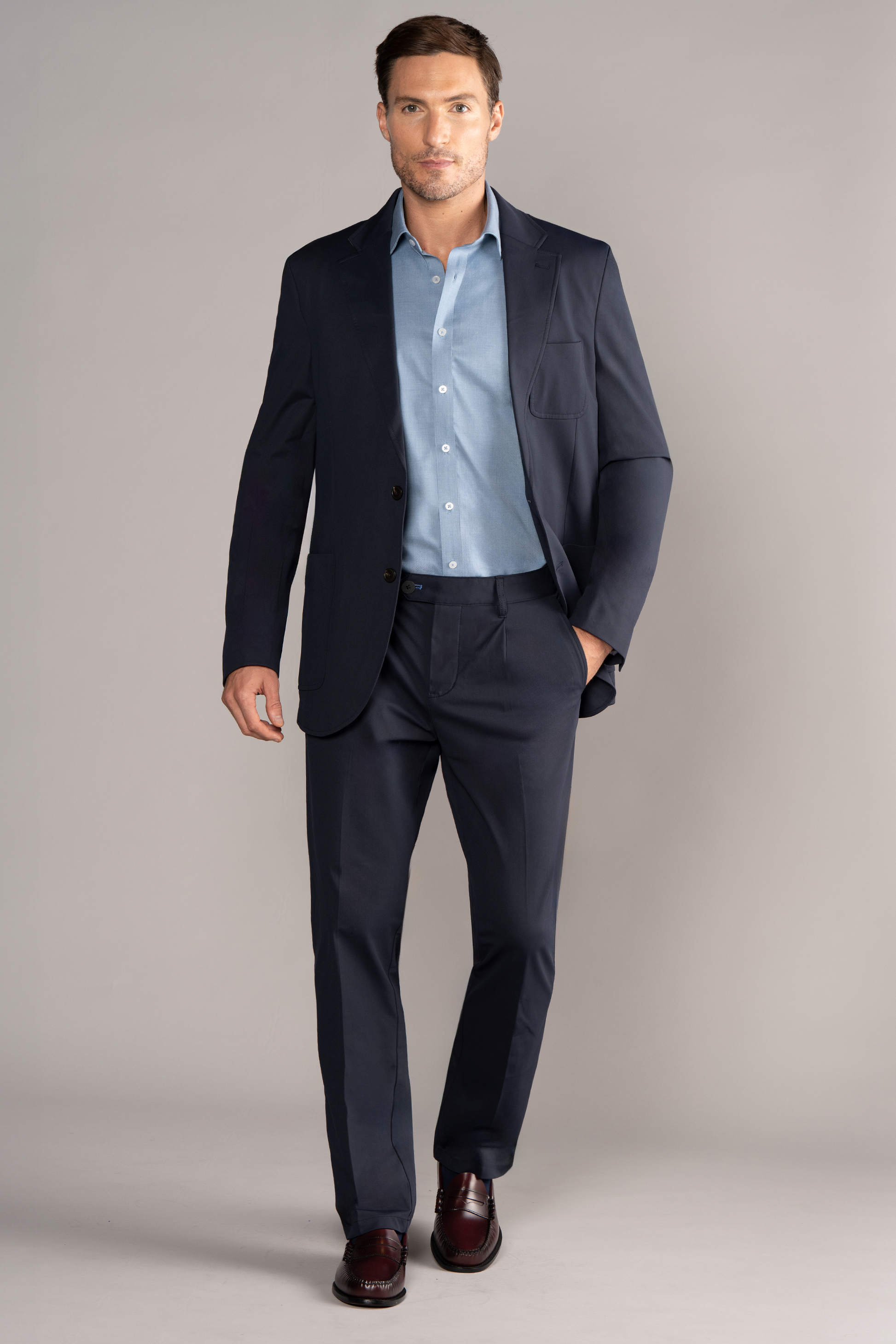 A man stands confidently against a neutral background, wearing Twillory’s Performance Patch Pocket Blazer in navy over a light blue dress shirt. He has one hand in his pocket, burgundy dress shoes, and a composed expression as he looks straight ahead.