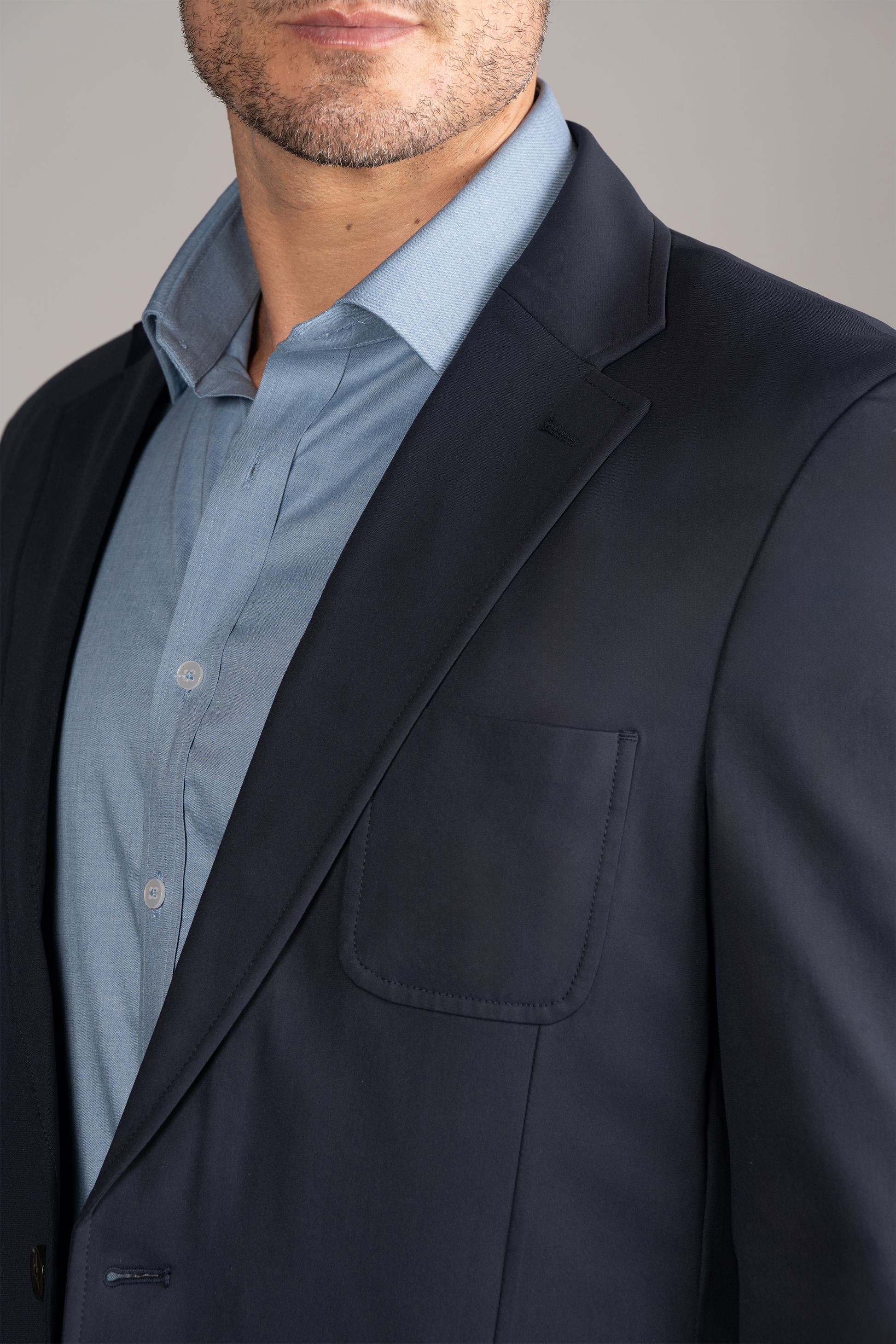 A man wears the Twillory Performance Patch Pocket Blazer in navy over a light blue dress shirt. The cropped image highlights the notched lapel and breast pocket, set against a plain light gray background.