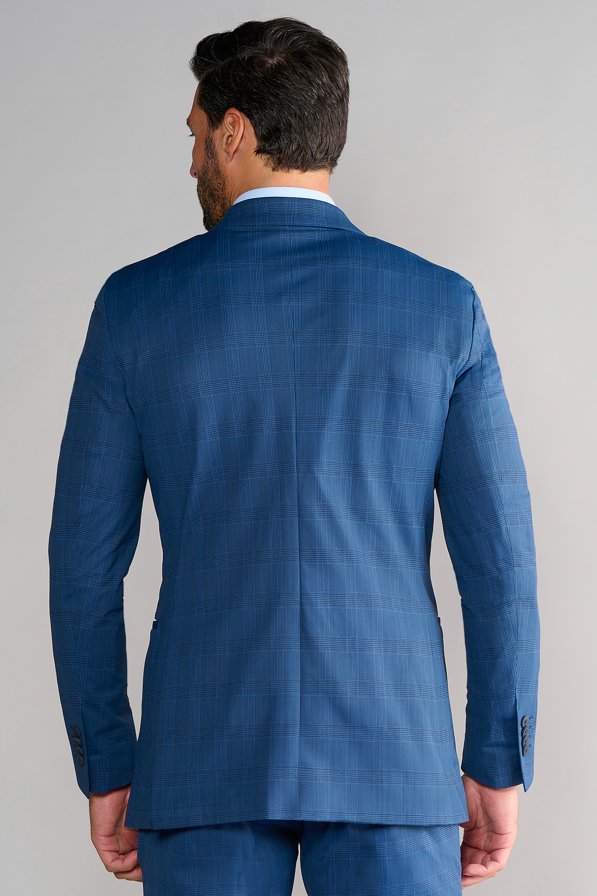 A man stands with his back to the camera, wearing the Twillory Performance Blazer in Blue Prince of Wales with matching trousers. His dark hair and beard are visible, arms at his sides, against a plain light-colored background.